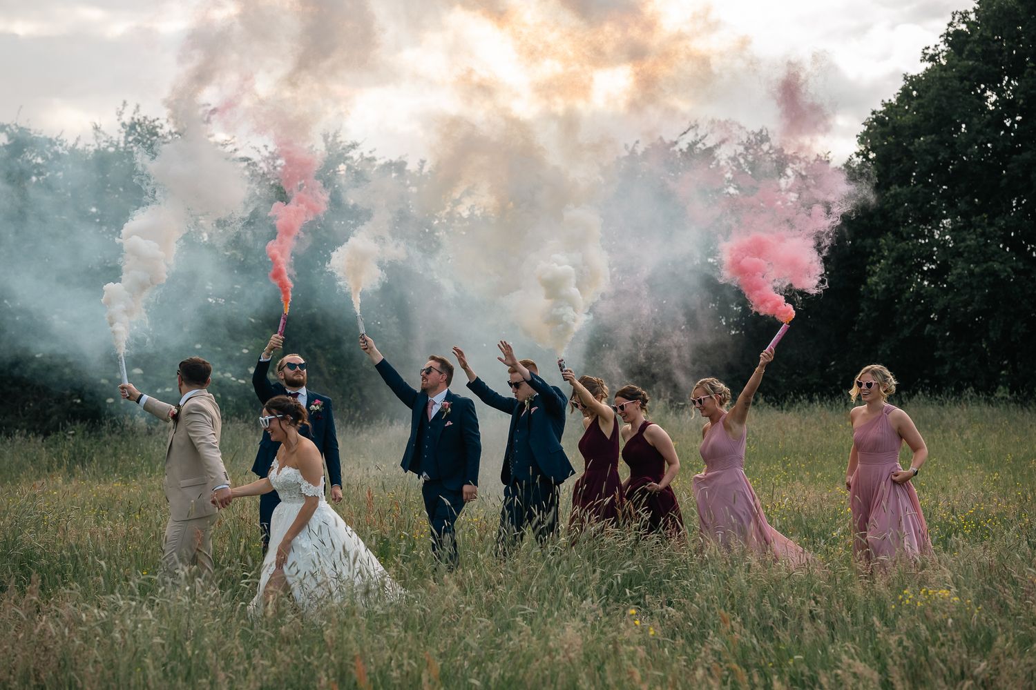 Bride, groom and bridal party walking through the field with pink and white smoke grenades at The Barn at Willerby