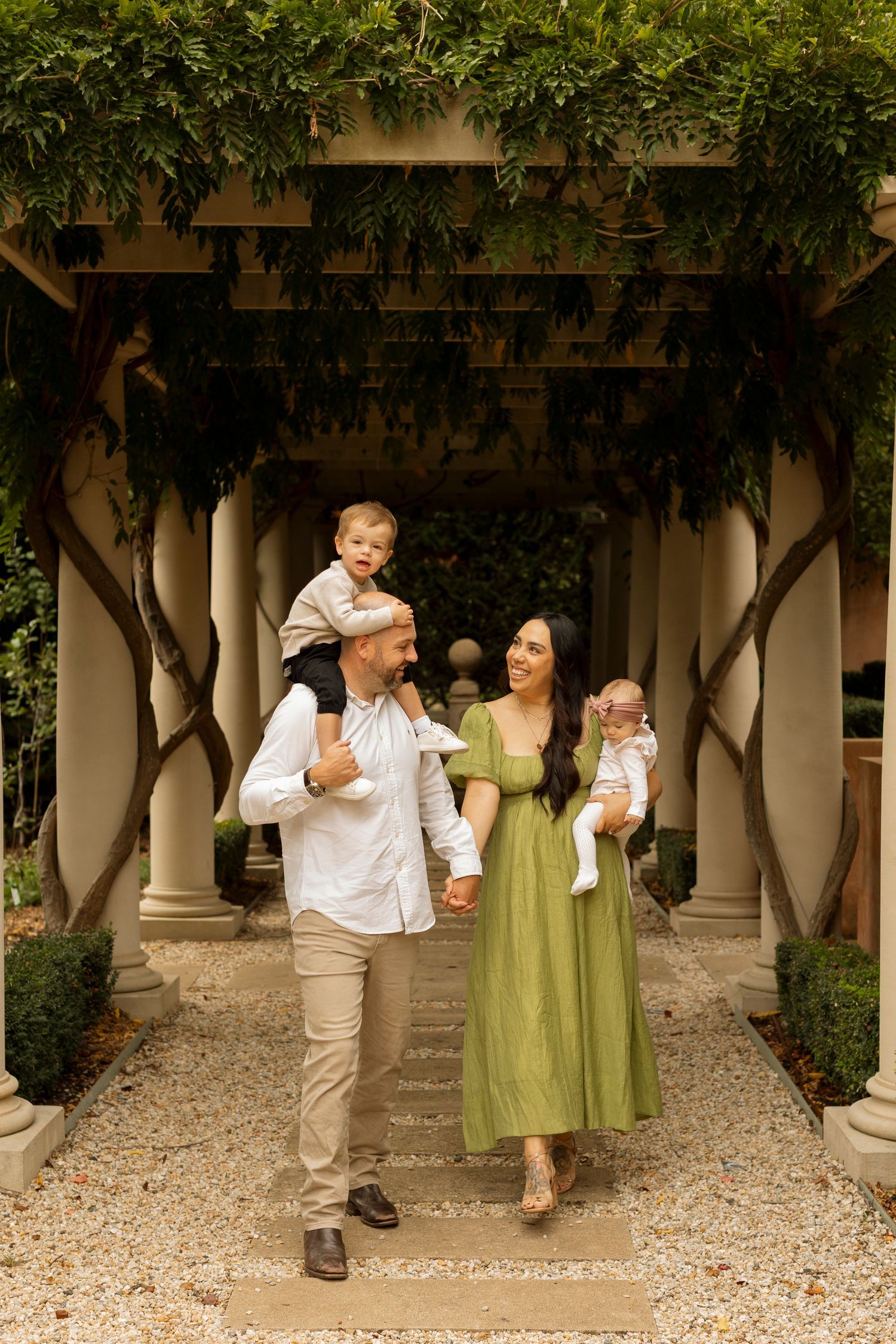 Three figures walking under a covered colonnade with stone pillars and greenery overhead.