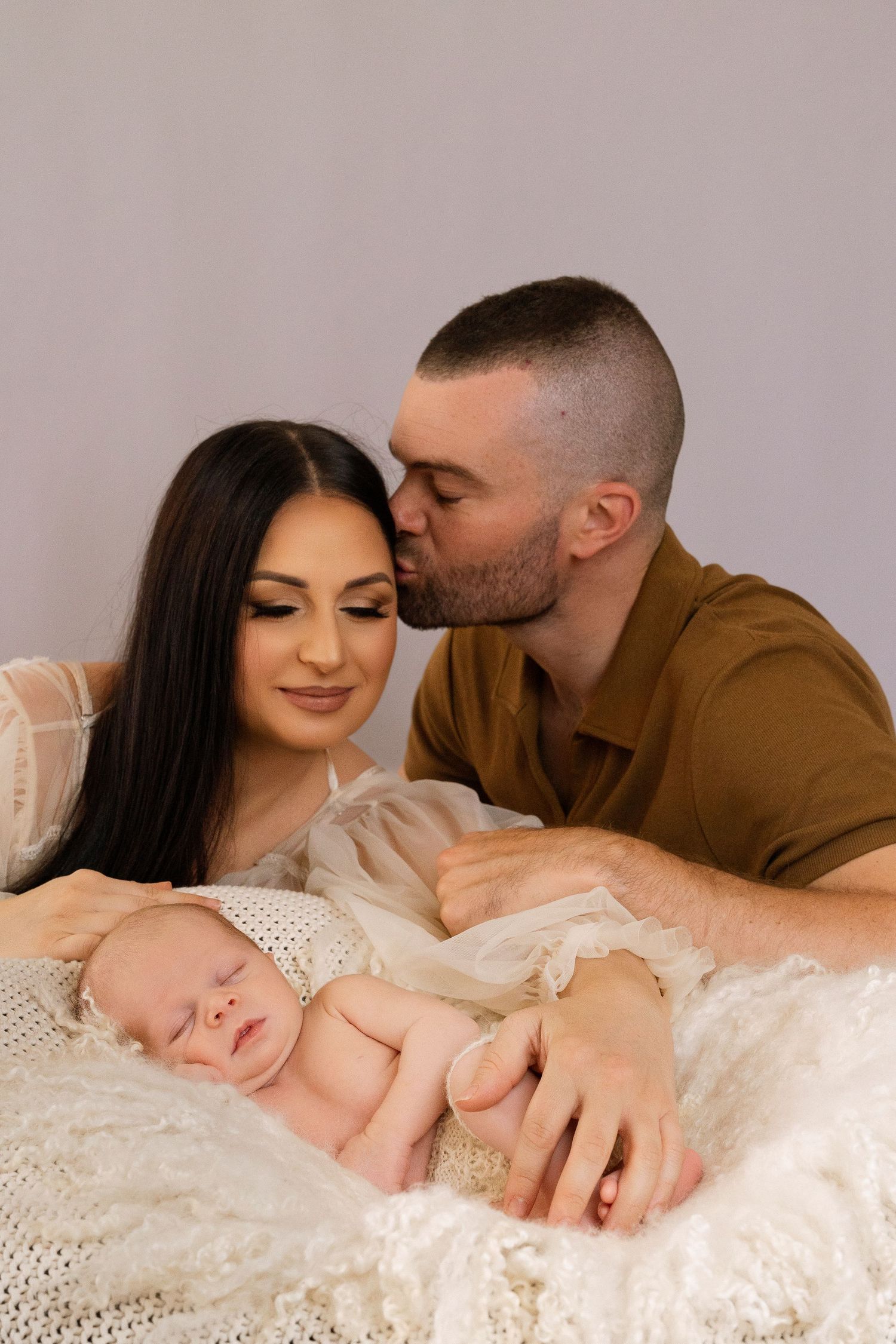 A sleeping newborn baby lies on white lace fabric while parents share an intimate moment above.
