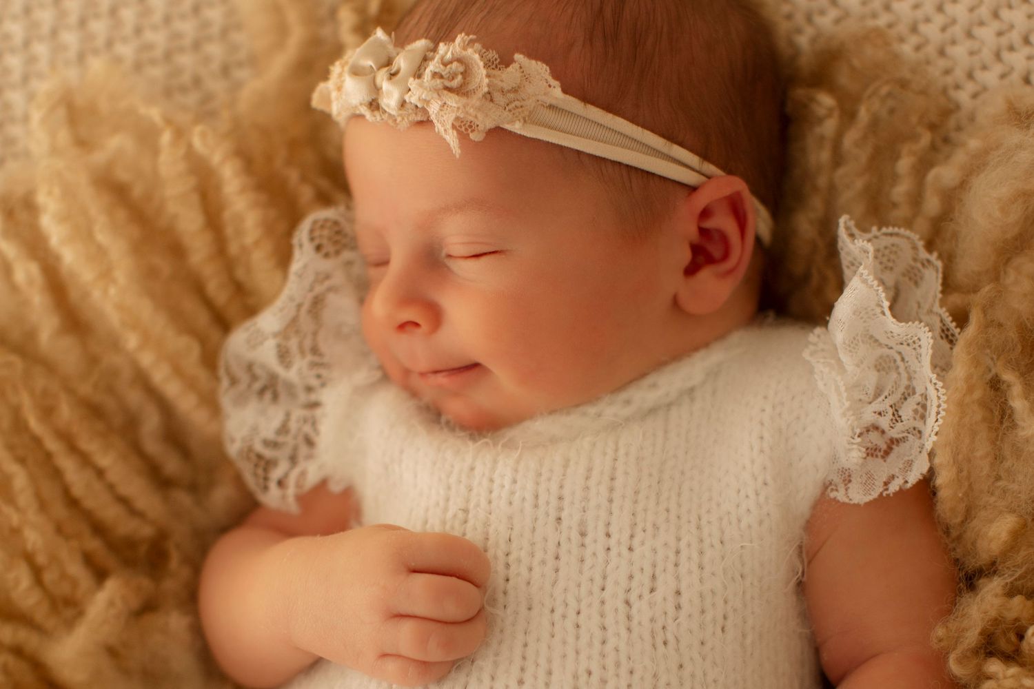 Newborn baby wearing white knit clothing rests peacefully on cream-colored blanket.