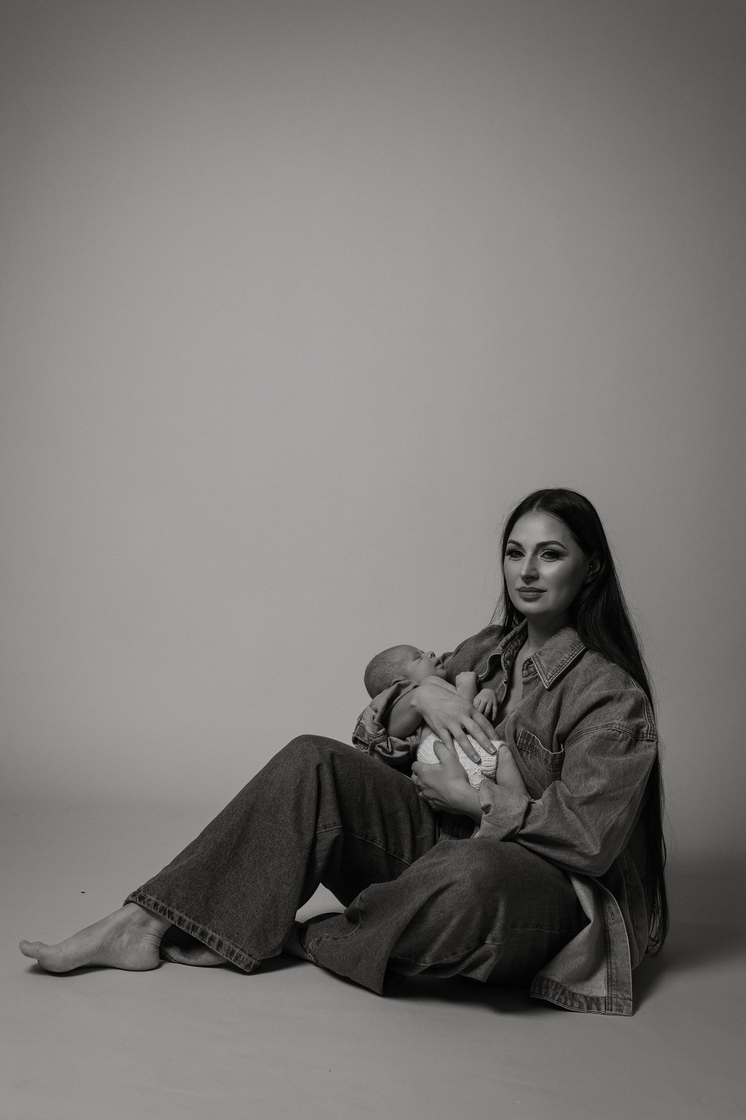 A person in denim clothing sits on the floor holding a sleeping infant in a minimalist black and white portrait.