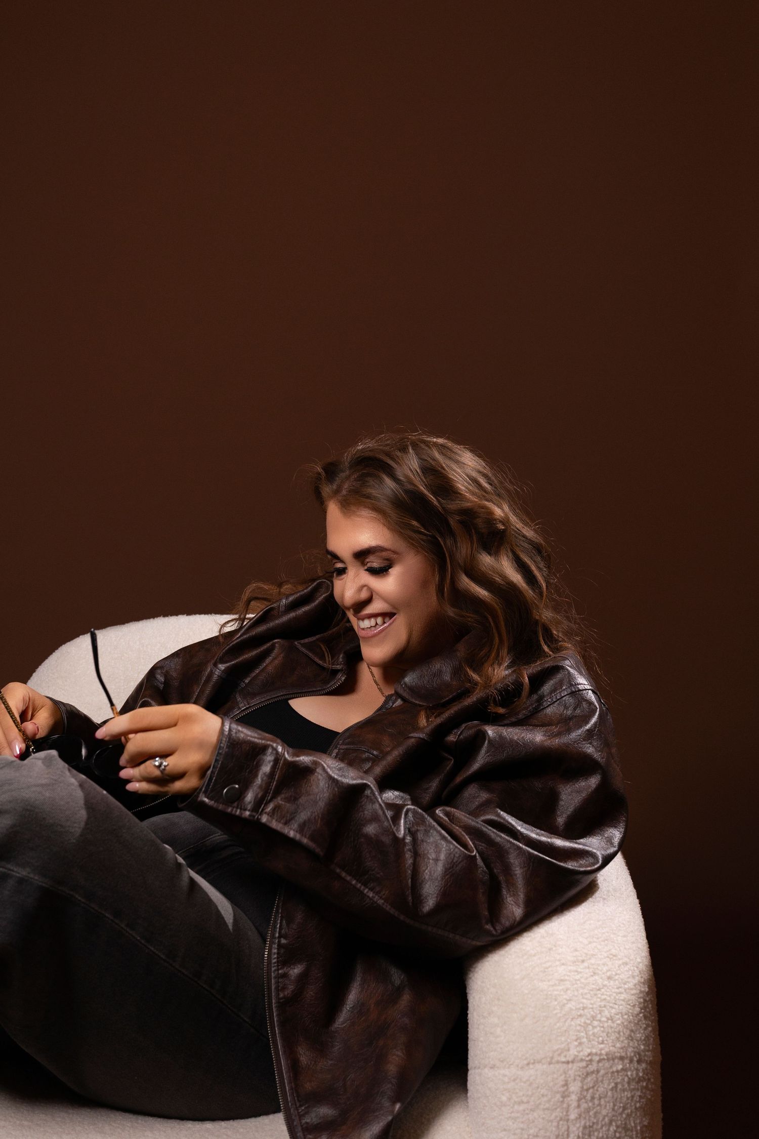 Smiling woman in brown leather jacket sitting relaxed in white chair against dark brown background.