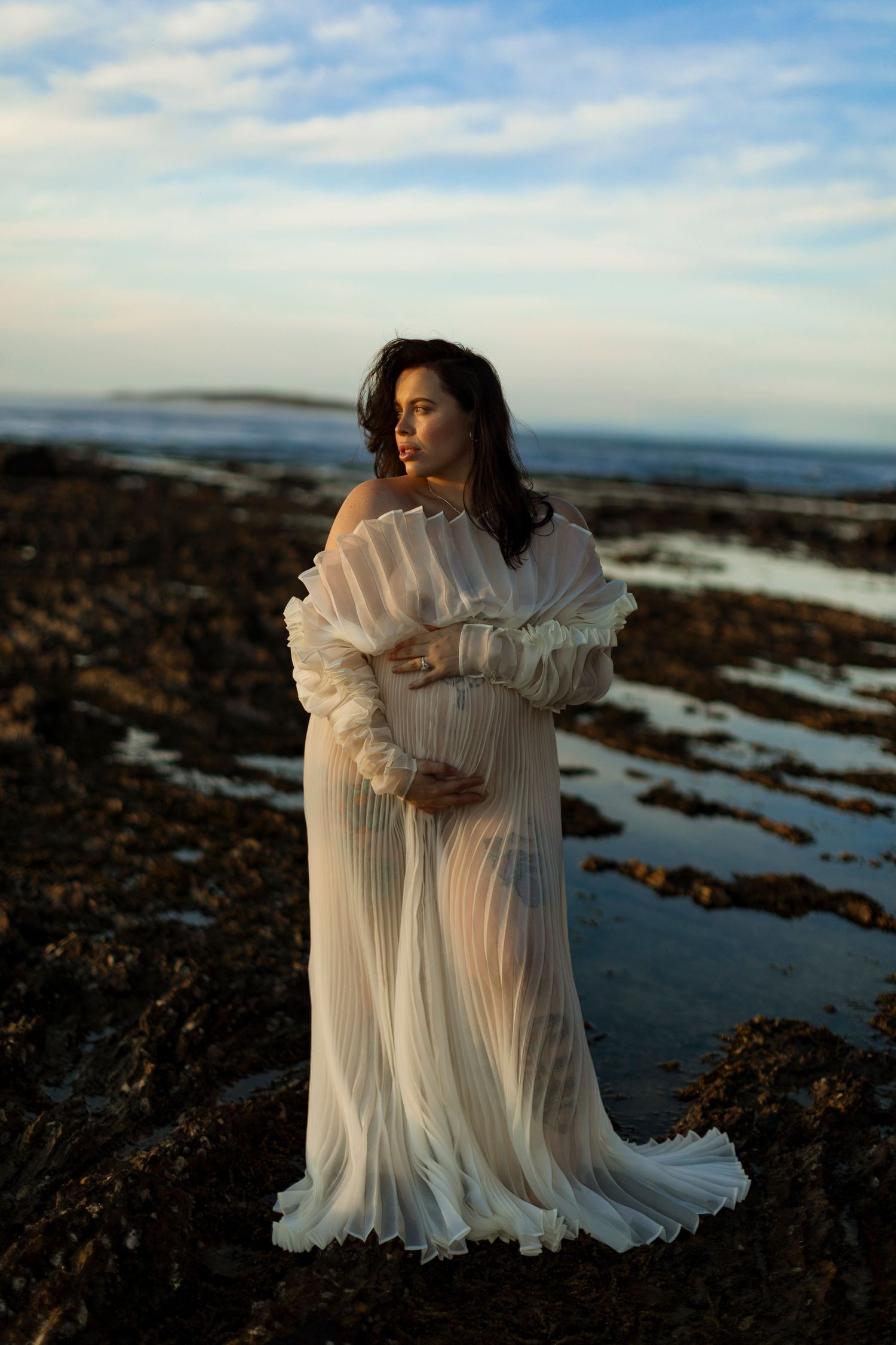 White flowing dress billows in coastal breeze on rocky shoreline.