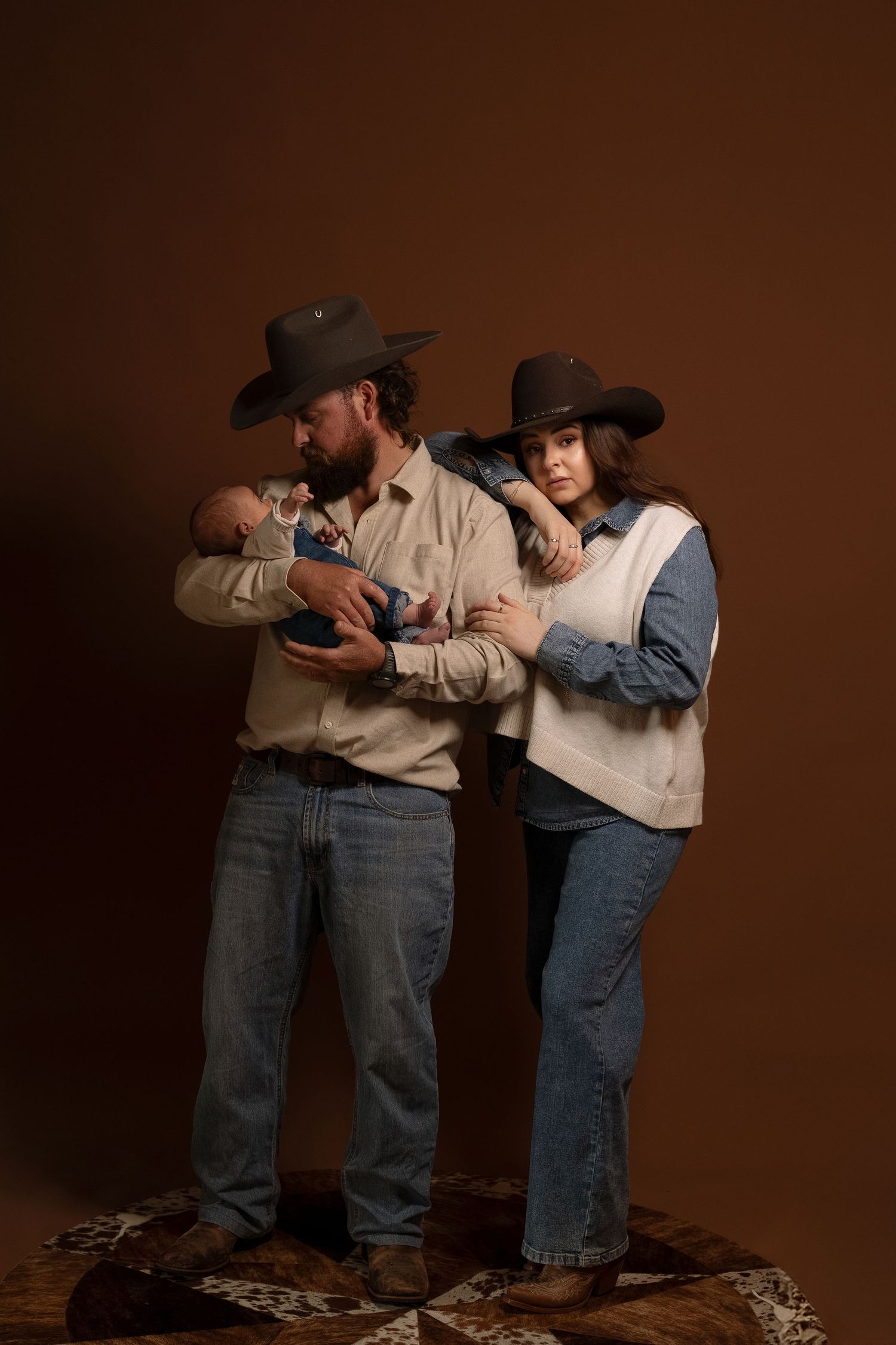 Couple in cowboy hats and jeans pose with newborn baby against brown studio backdrop.