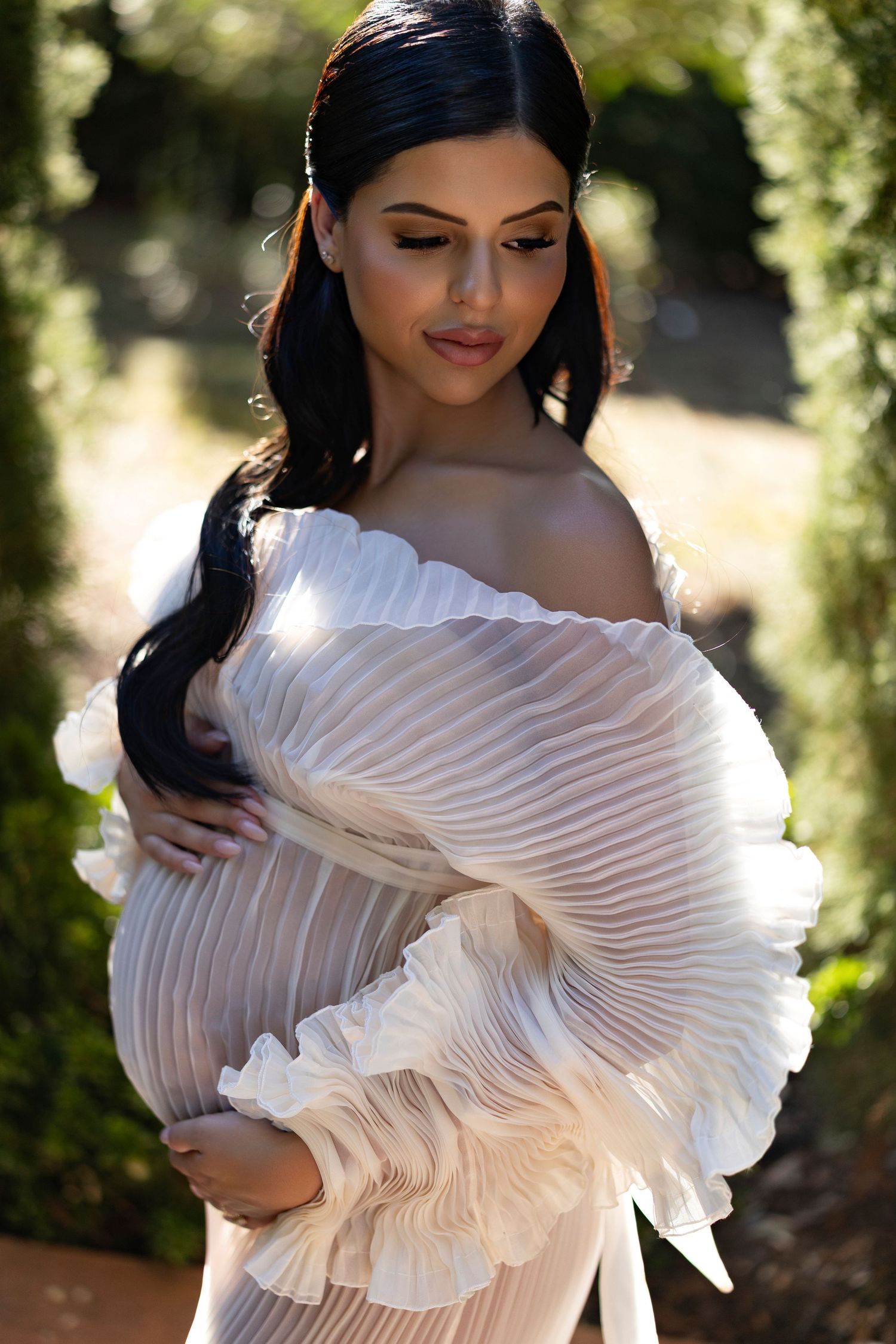 A figure in a white pleated ruffled dress poses elegantly in a sunlit outdoor setting.