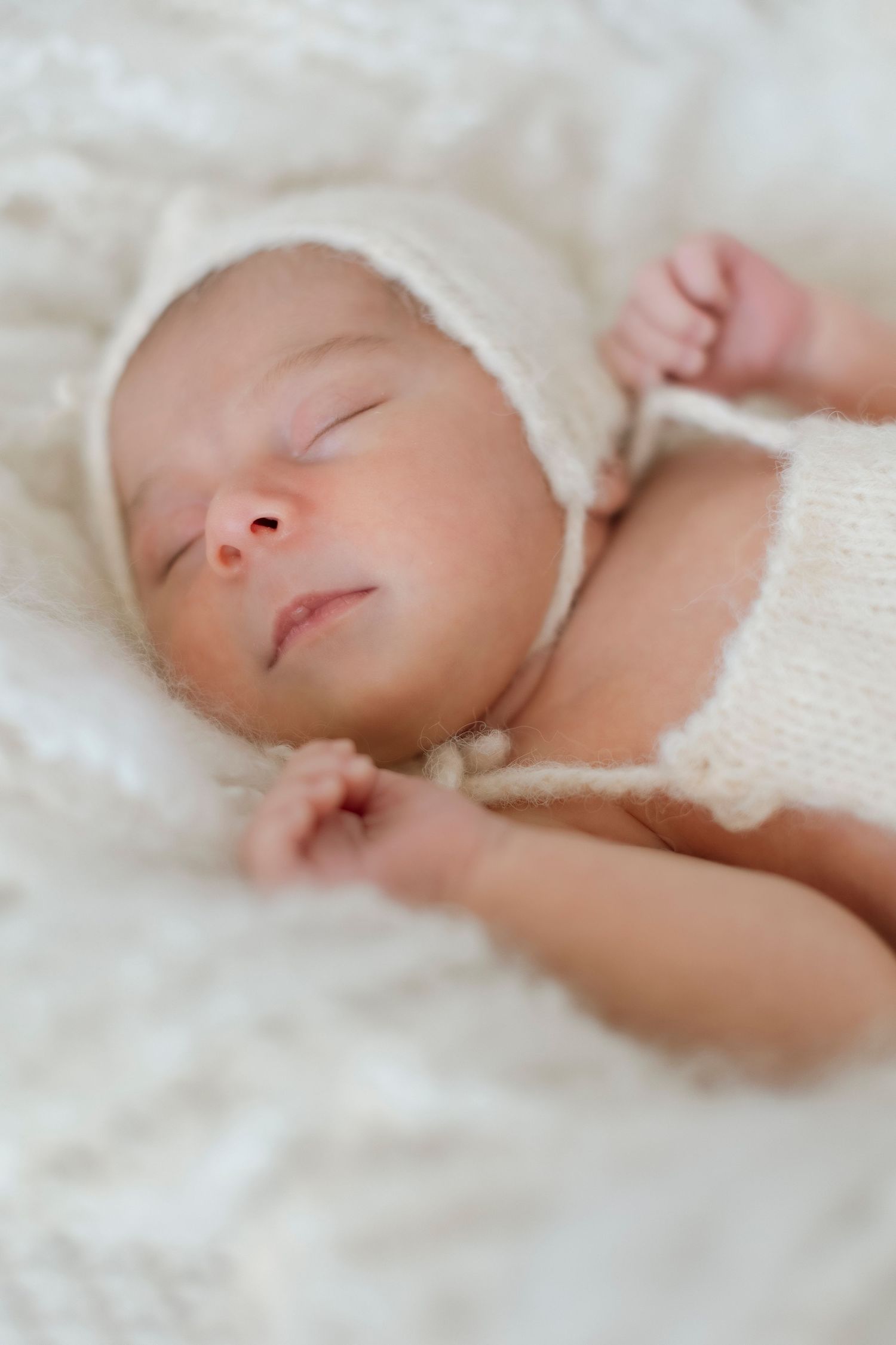 A peaceful sleeping newborn wearing a soft white knit bonnet rests on white bedding.