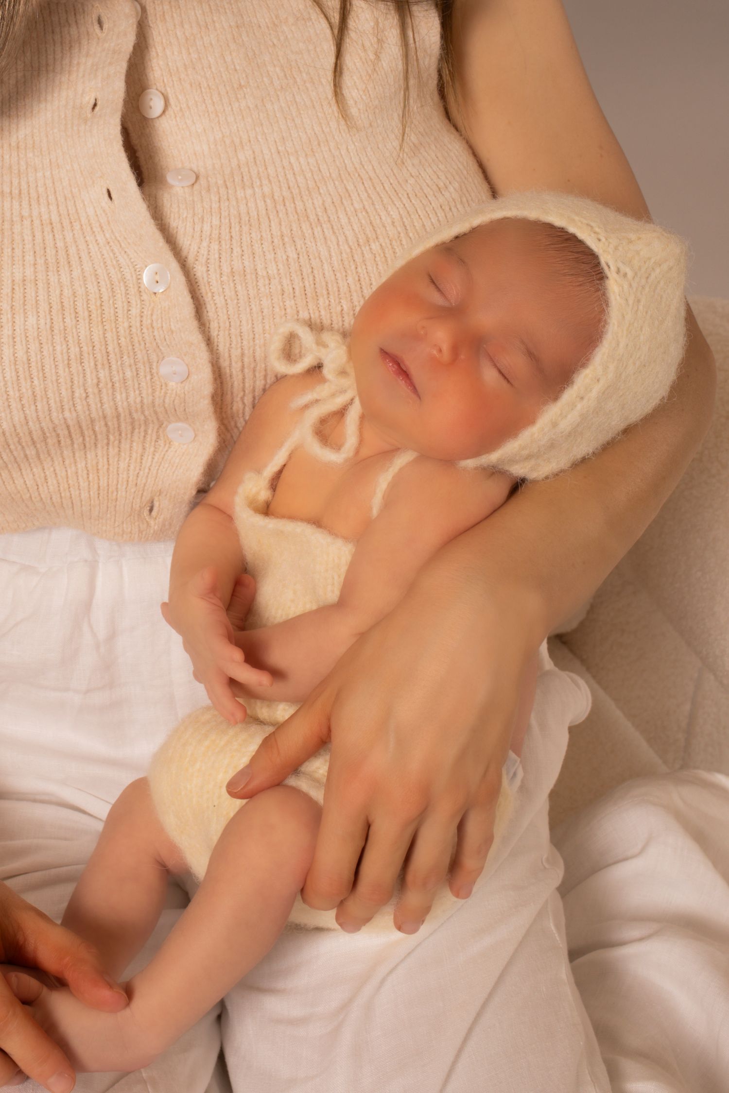 A sleeping newborn baby wearing a cream-colored cardigan lies peacefully on white bedding.