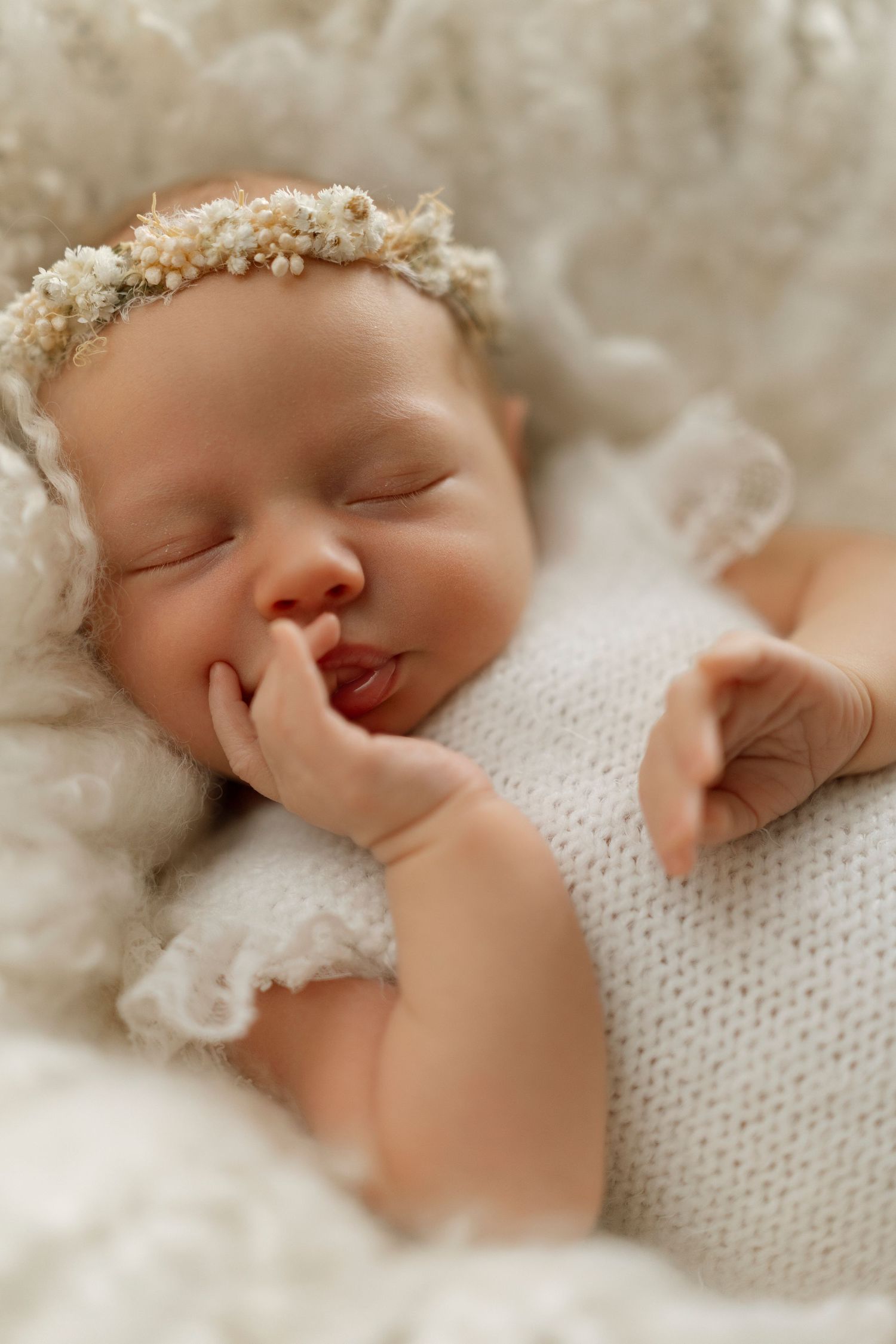 A sleeping baby wearing a delicate white knit outfit and floral crown rests peacefully on soft white fabric.