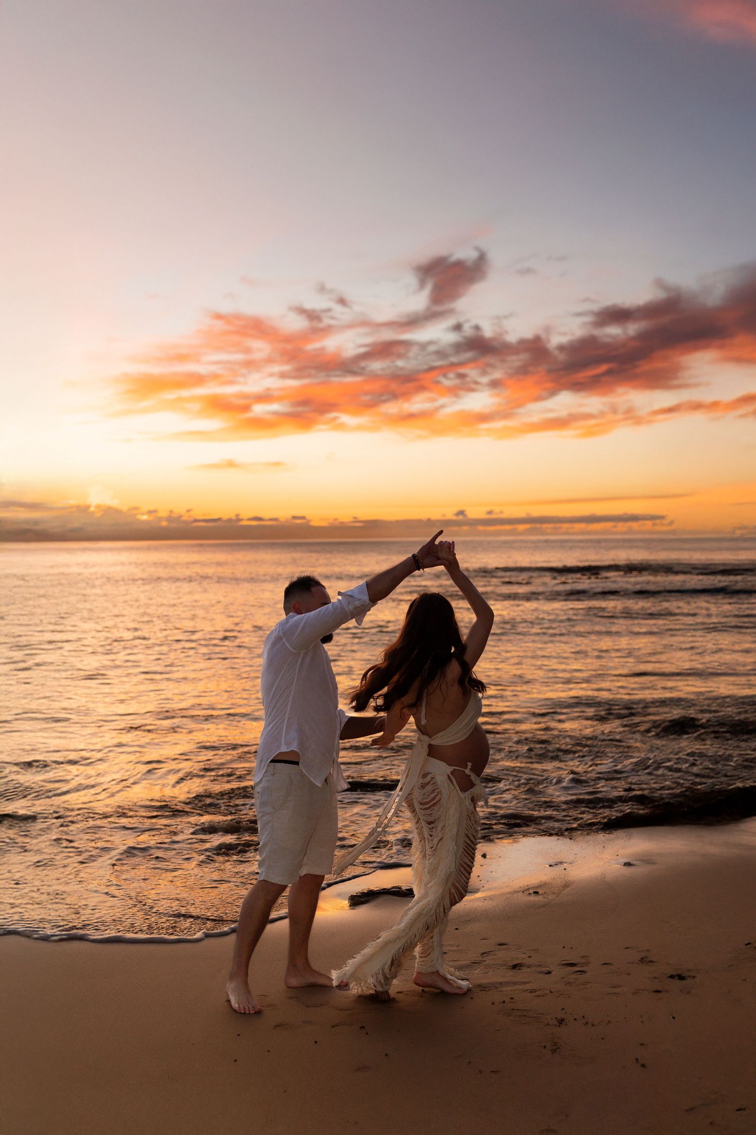 Two people dance together on a beach at sunset with vibrant orange and pink clouds reflecting on the ocean water.