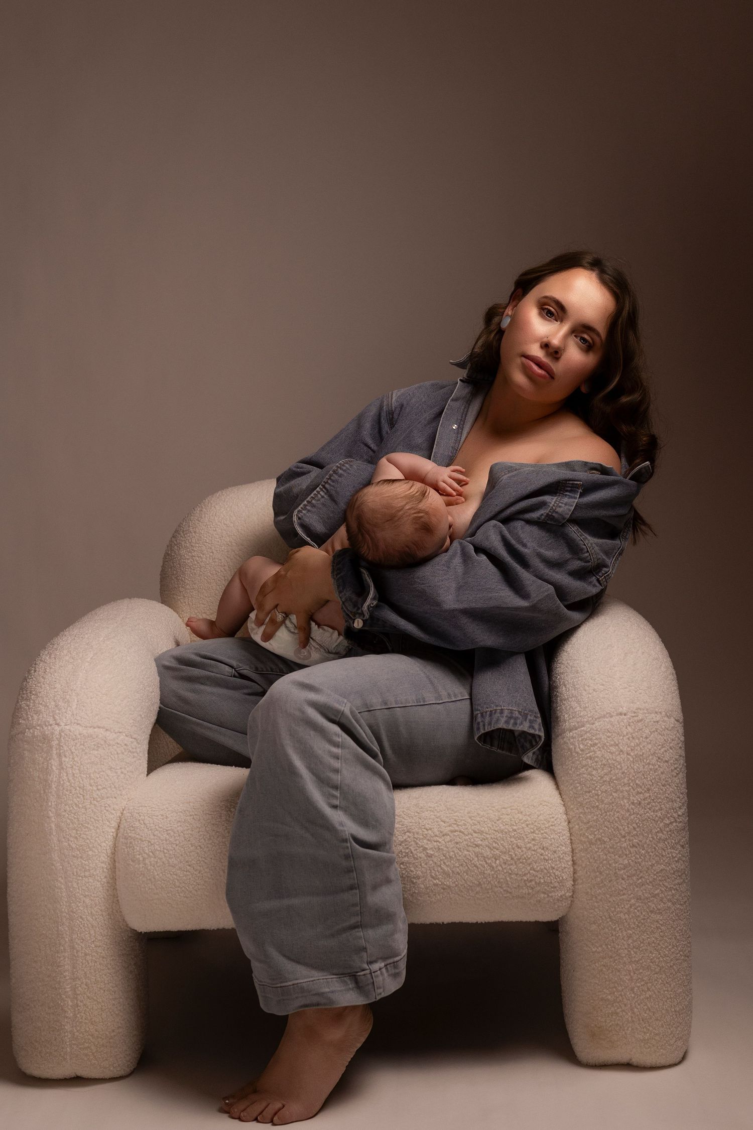 A figure in a gray dress lounges on a curved white textured sofa in a studio setting with moody lighting.