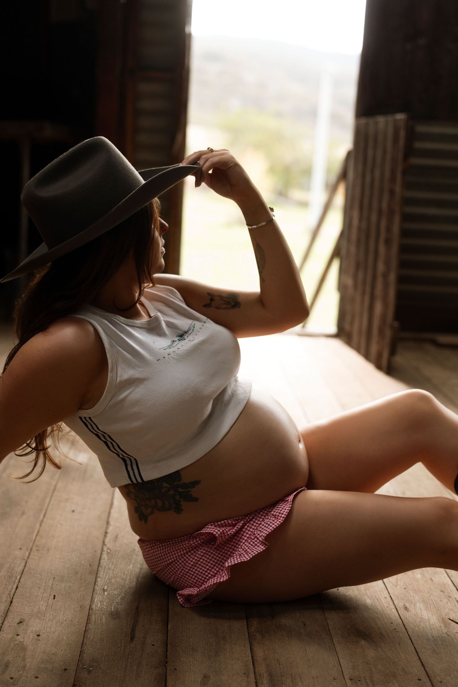 Pregnant woman in white crop top, pink shorts, and gray cowboy hat sitting on wooden floor in rustic barn.