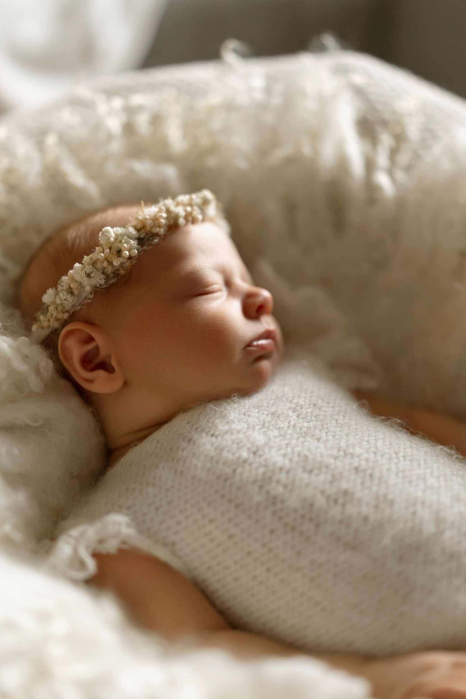 Peaceful newborn baby wearing white knit wrap and floral crown resting on soft bedding.