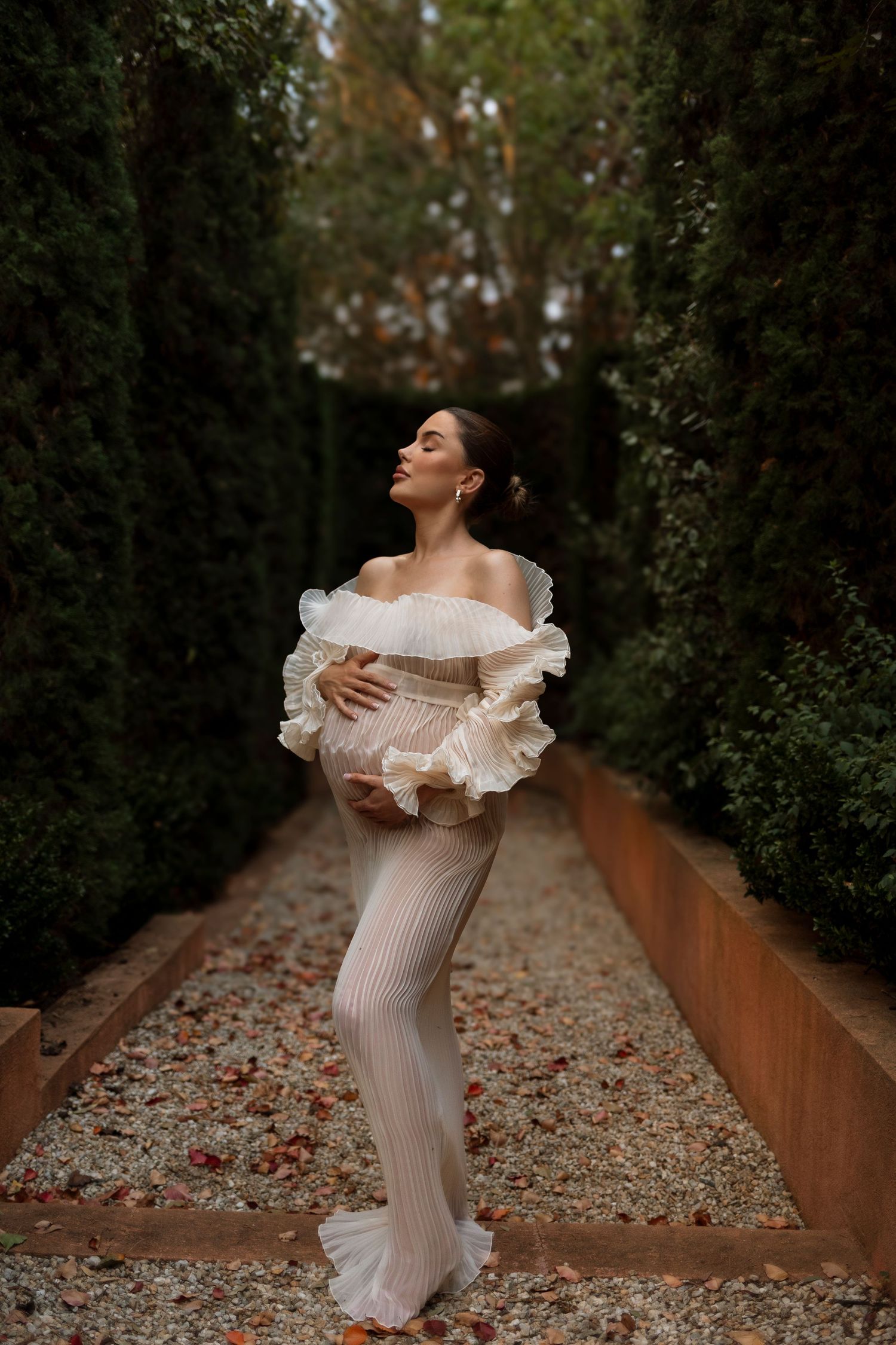 Maternity portrait in garden path wearing flowing white dress with ruffled sleeves.