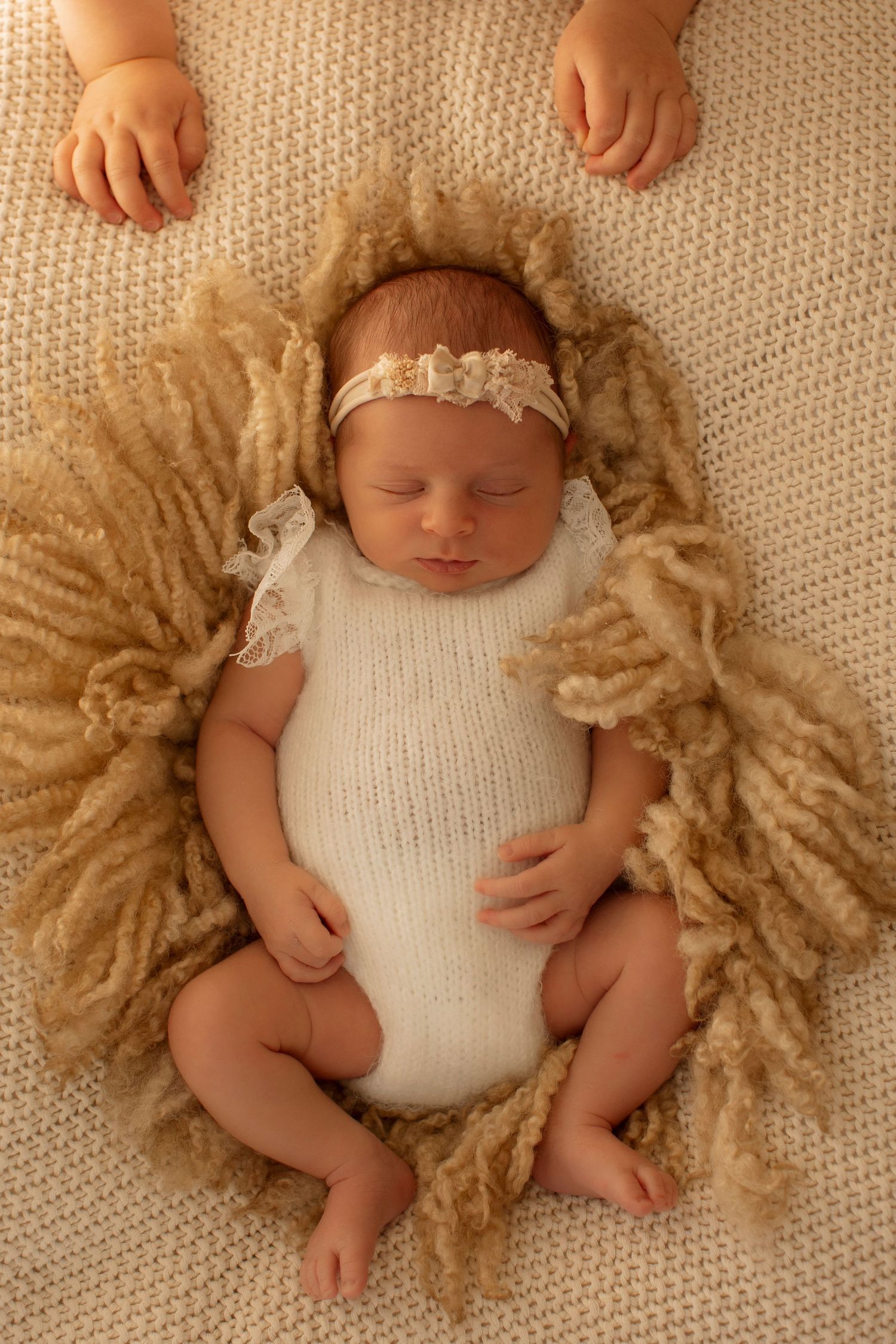 Sleeping newborn wearing white knit romper and lace headband lying on beige textured blanket in studio setting.