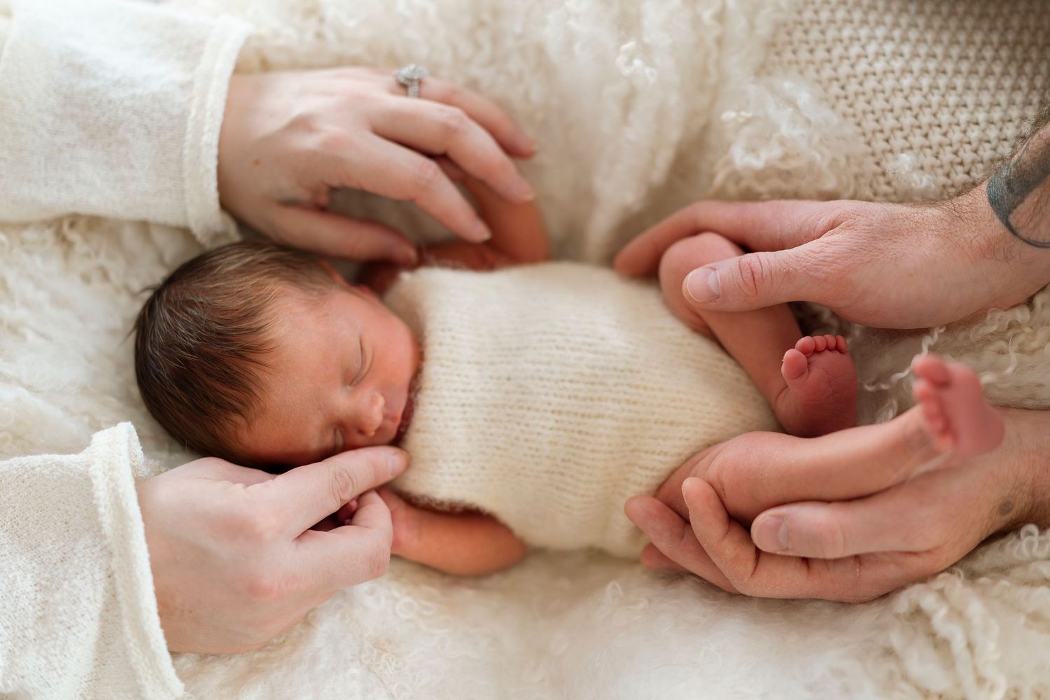 A sleeping baby wrapped in a cream colored blanket rests peacefully while being gently touched by adult hands.