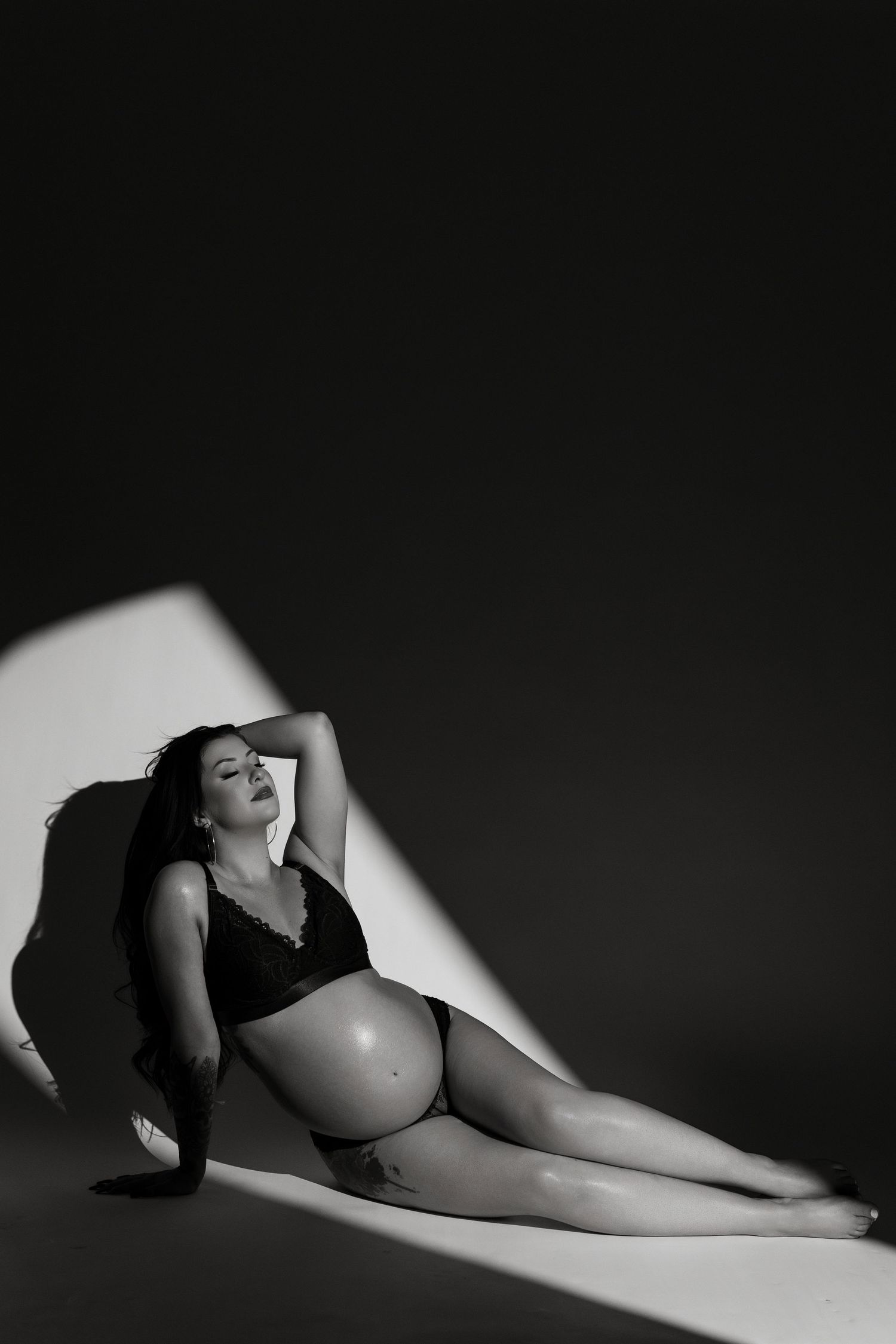 Black and white maternity photo of an expectant mother in lingerie reclining in dramatic lighting.