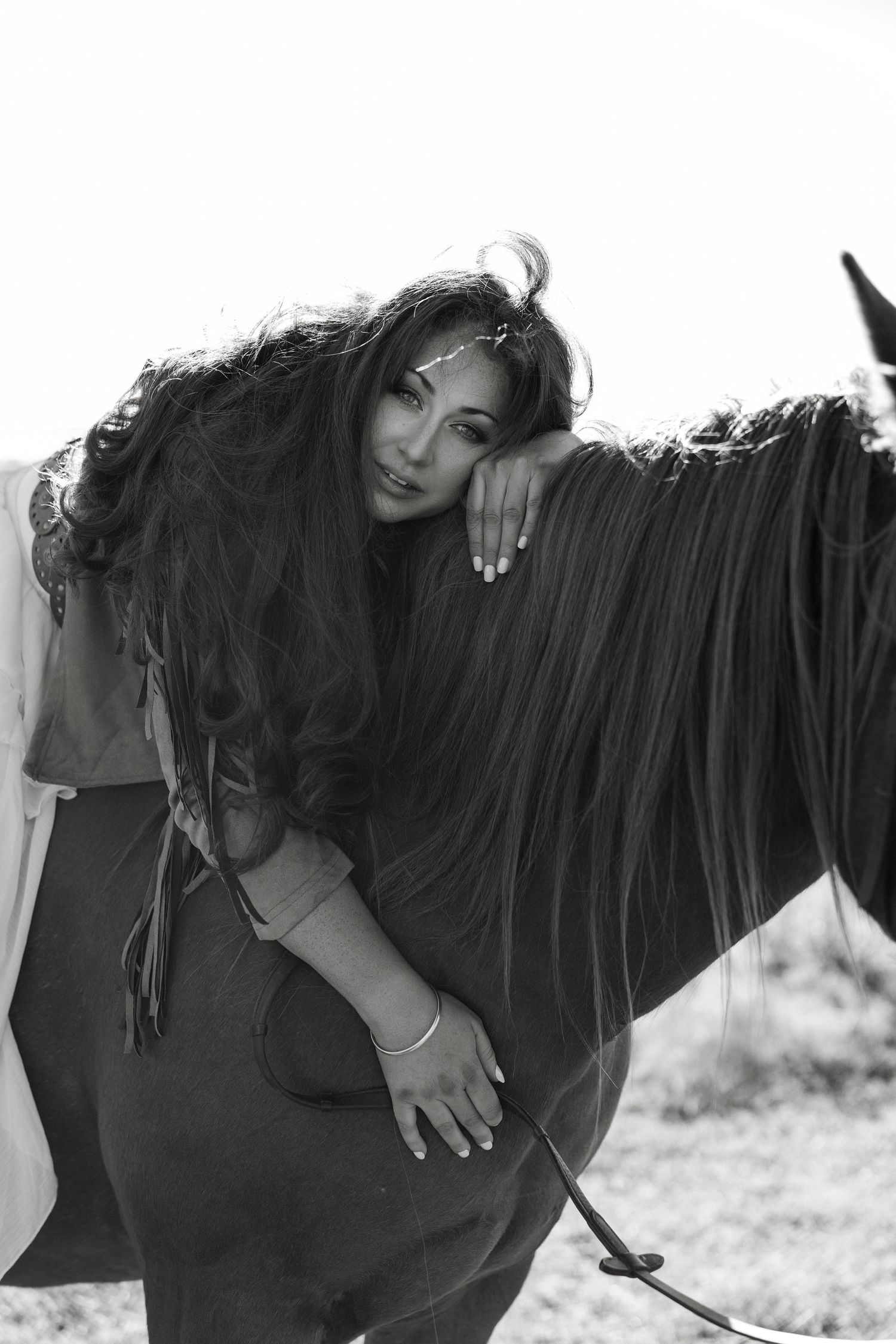 Black and white portrait of a person with long dark hair leaning against a horse's neck in an artistic pose.