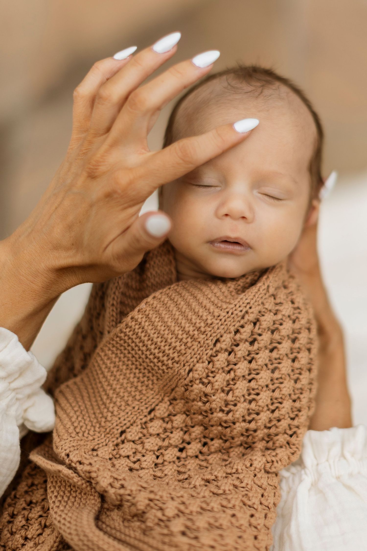 Sleeping baby wrapped in intricate brown knit blanket with gentle hands caressing.