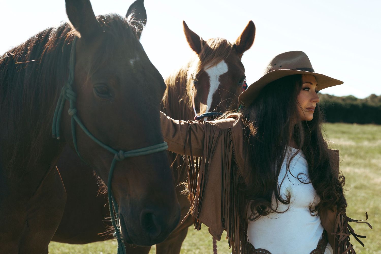 A woman in a cowboy hat stands with two horses in a sunny field, one dark brown and one with a white face.
