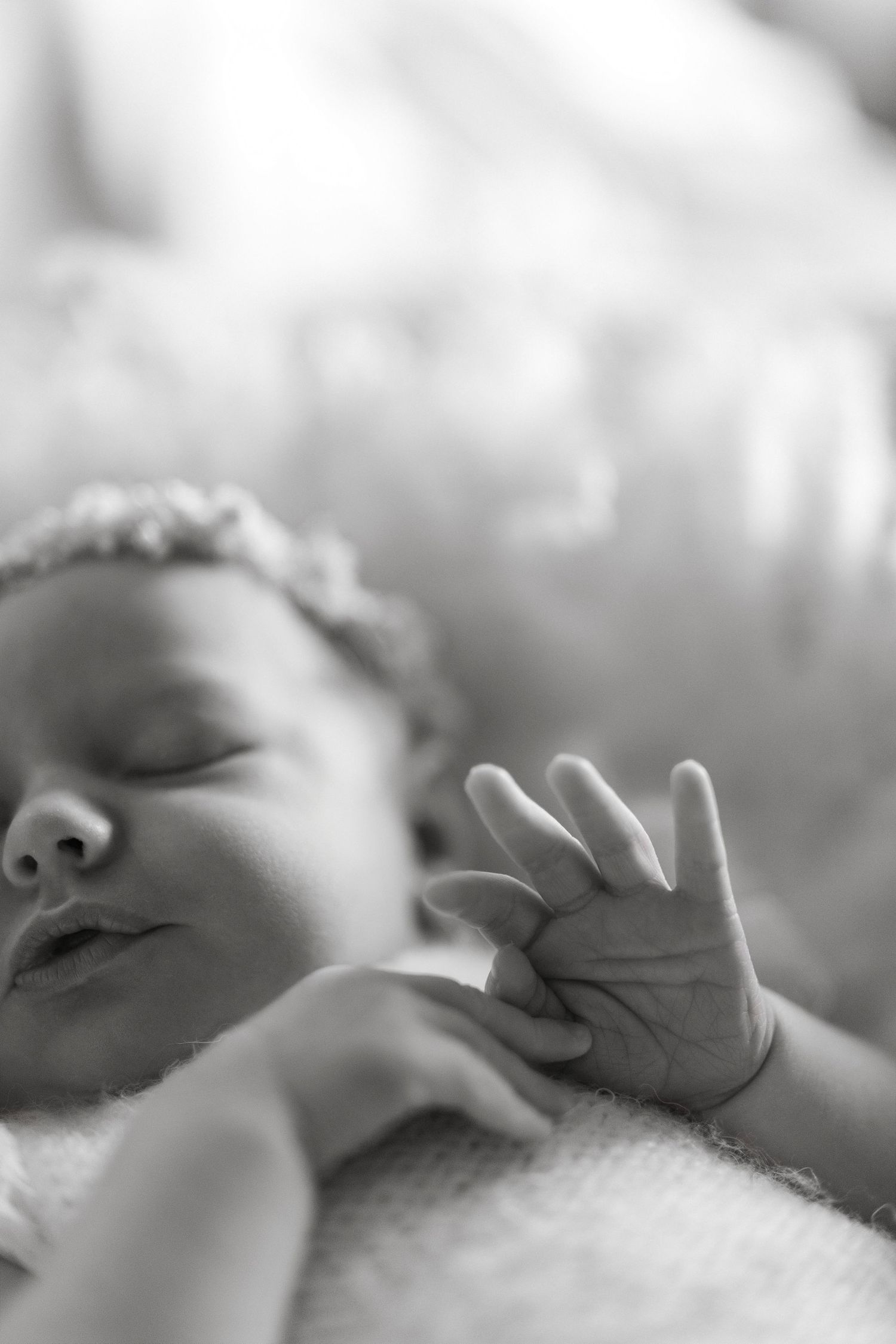 Close-up black and white image of a sleeping newborn baby's face and tiny hand against soft bedding.