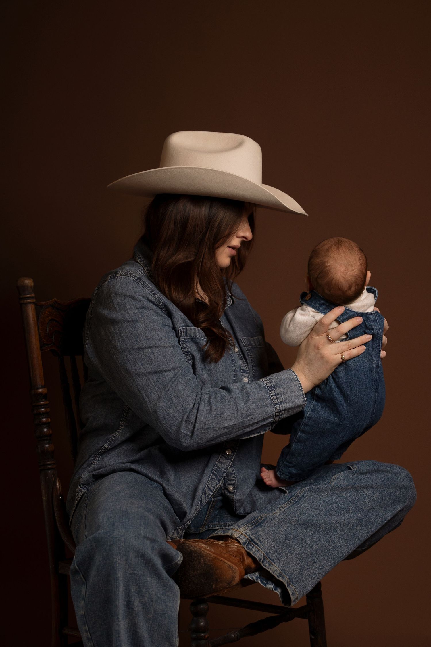Woman in cowboy hat and denim outfit sitting in wooden chair holding newborn baby in studio portrait.