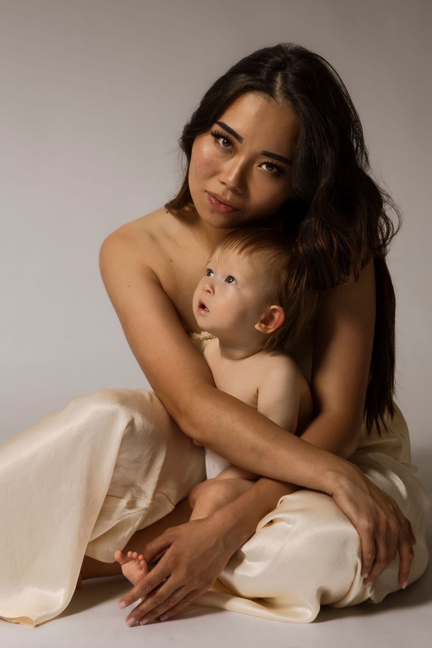 Intimate studio portrait of mother and baby wrapped in cream-colored fabric.