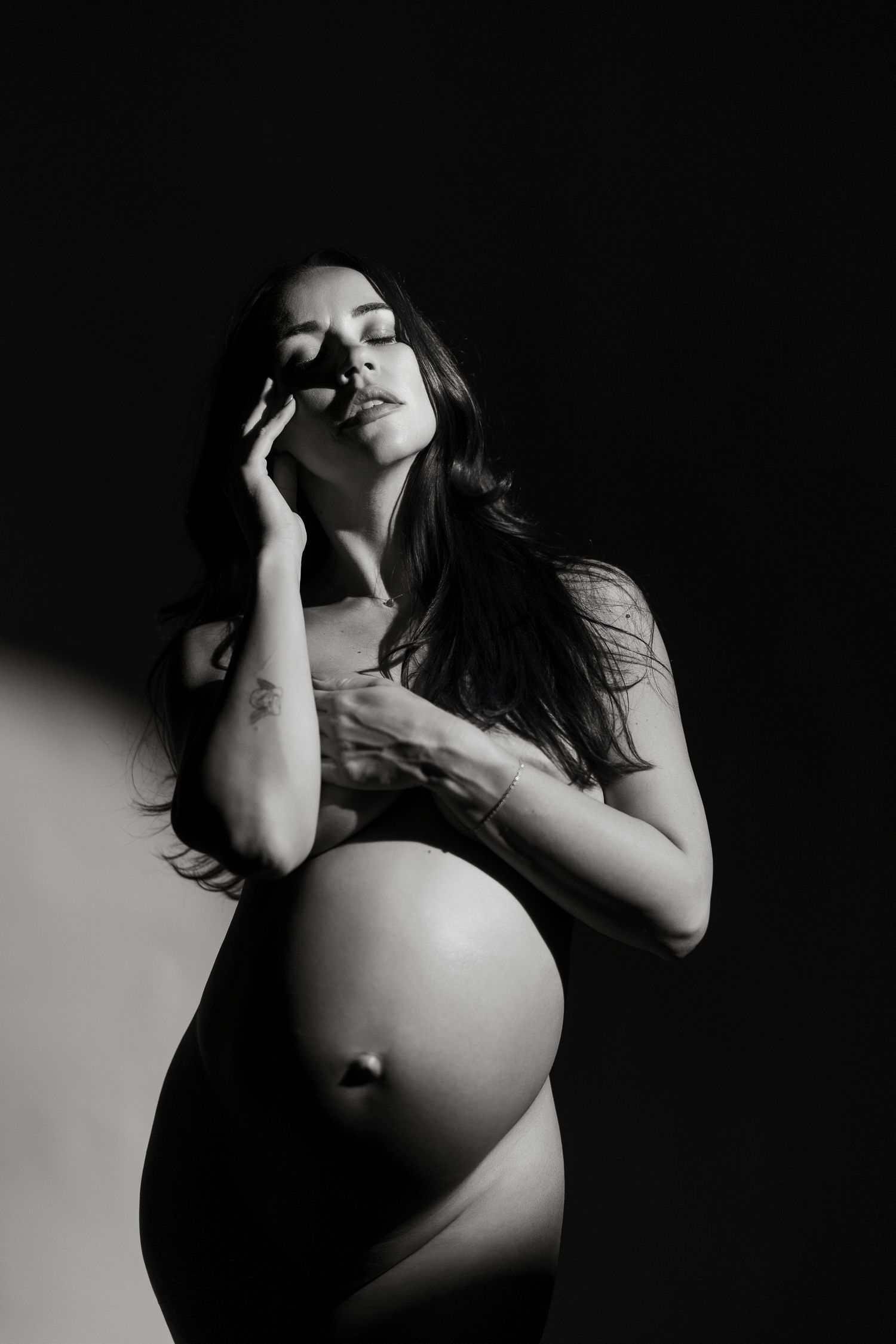 Black and white maternity photo of a pregnant woman posing gracefully against a dark background.