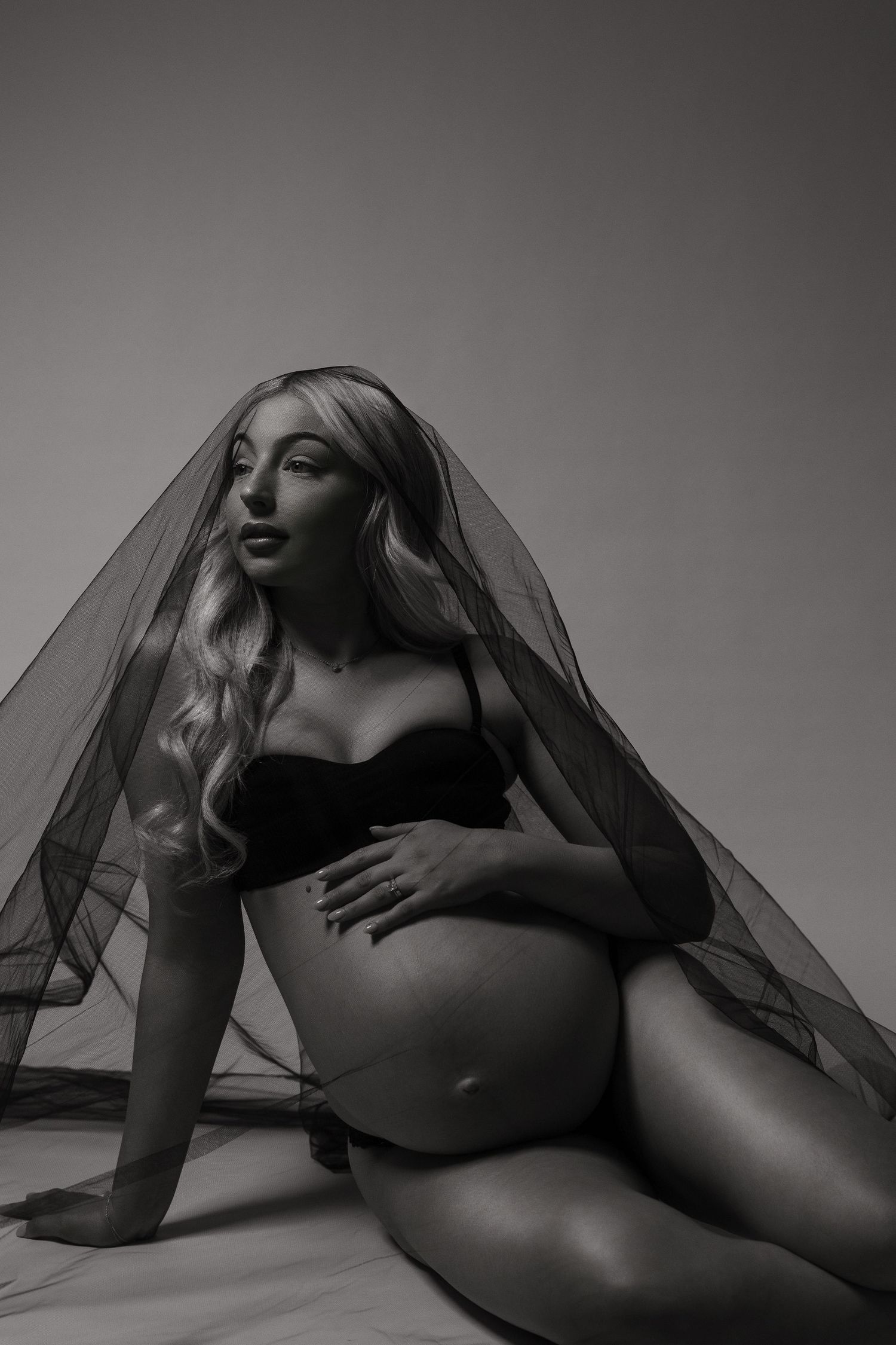 Black and white maternity photo of a blonde woman in a bra under sheer fabric, cradling her baby bump while seated.