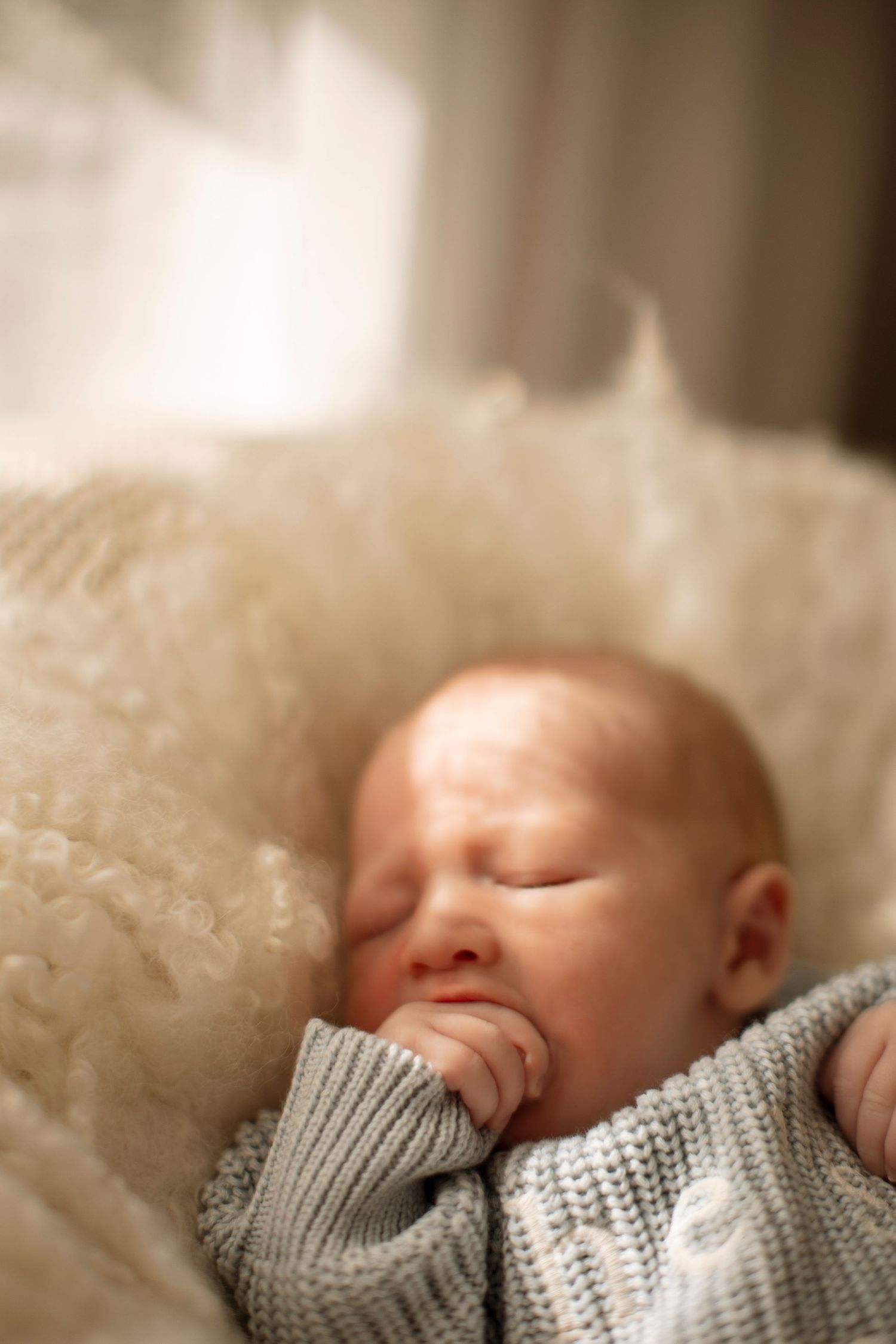 A sleeping baby wearing a cozy gray knit sweater rests peacefully on soft cream-colored bedding.