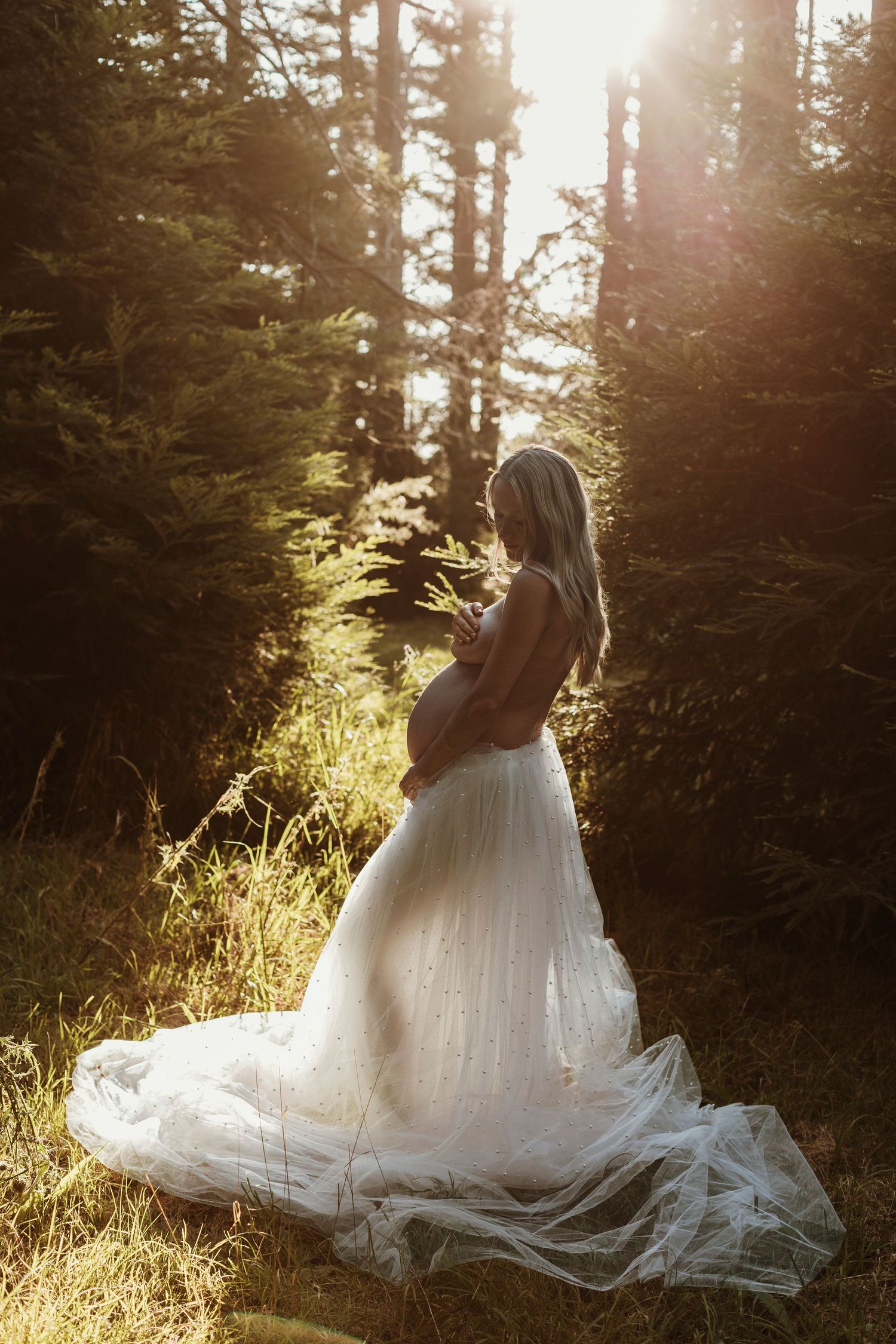 Dreamy portrait in white flowing dress among tall pine trees at golden hour.