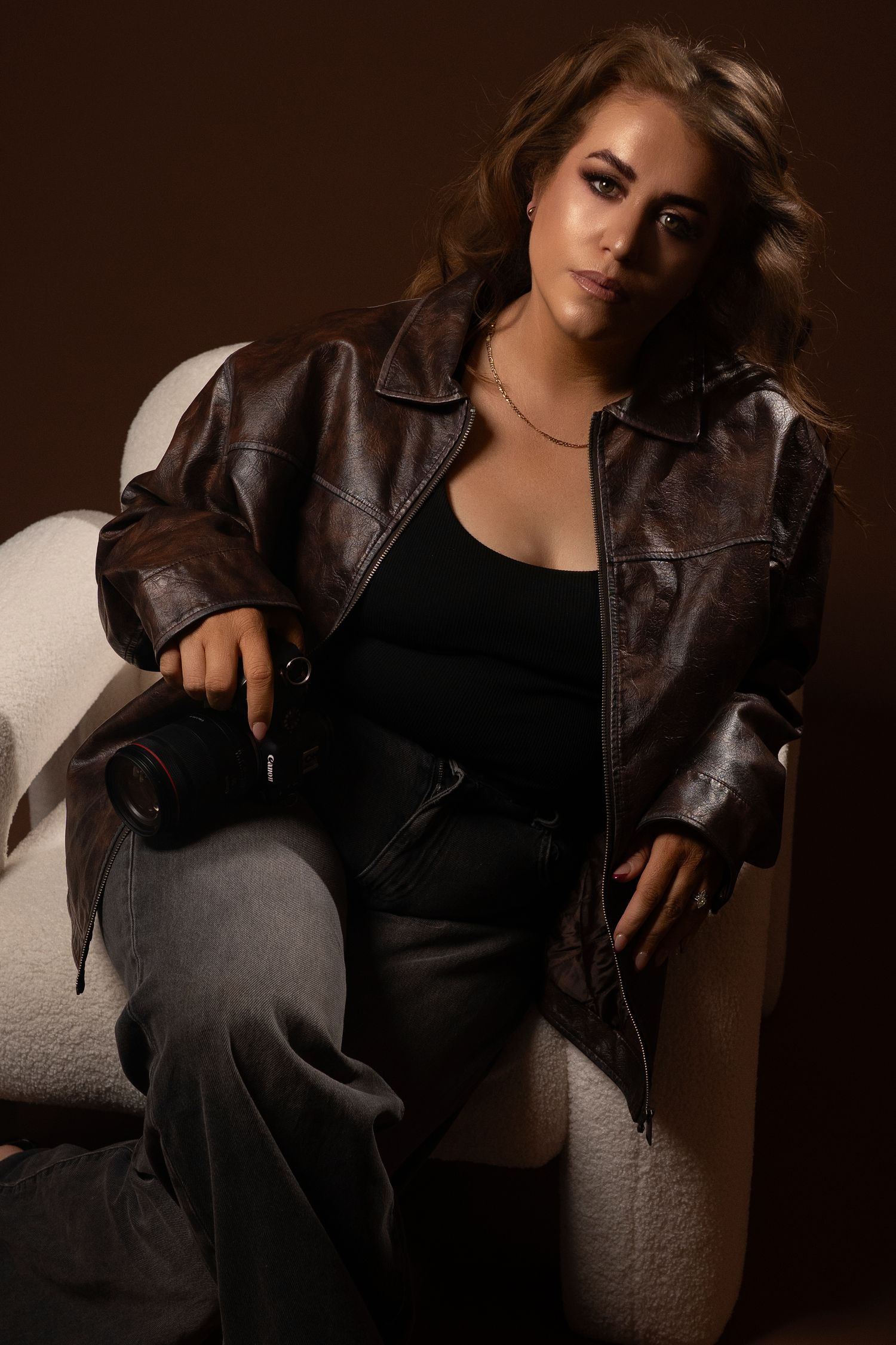 Model wearing a brown leather jacket and black top poses against dark background in dramatic lighting.