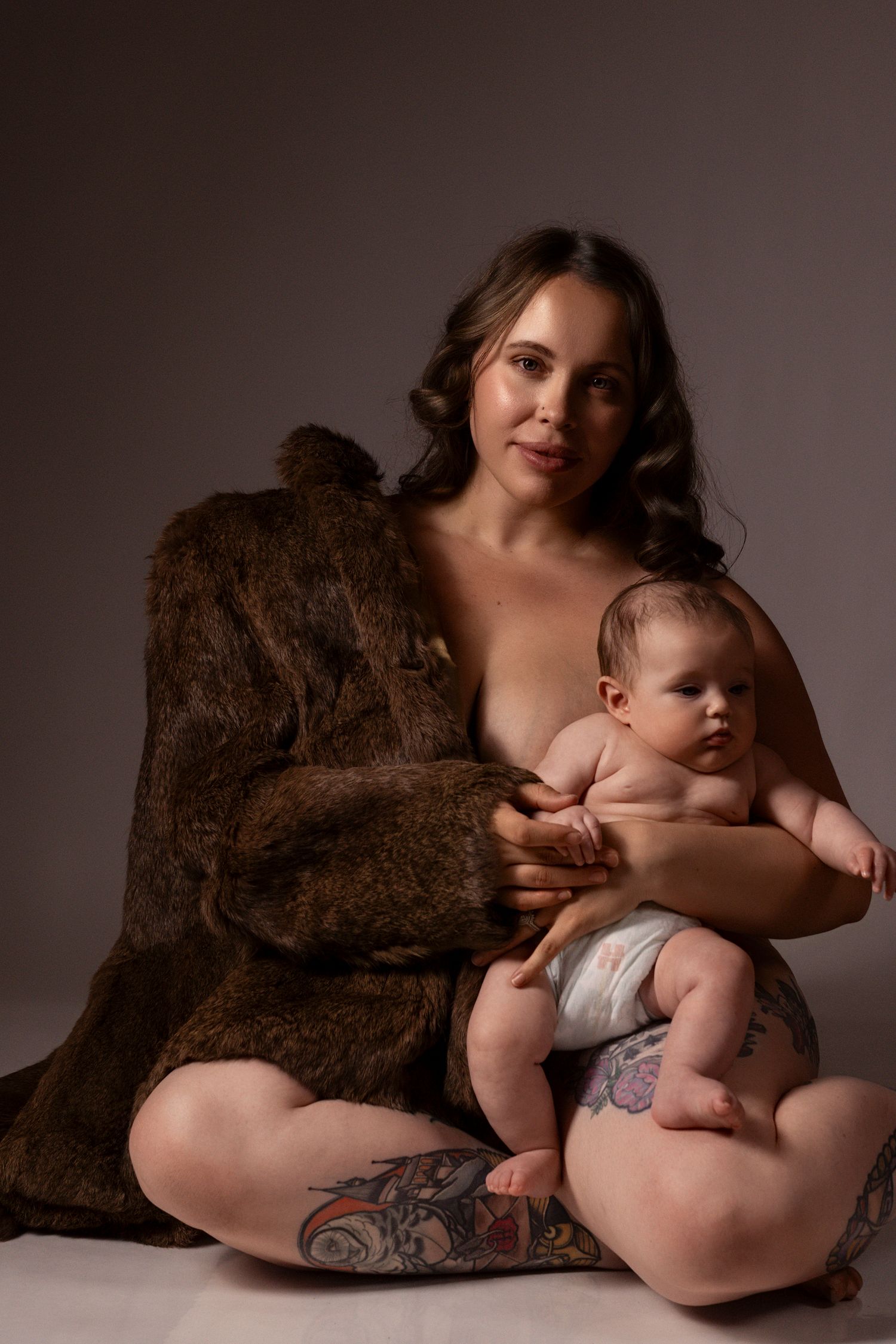 A tattooed mother in a fur coat sits cross-legged while breastfeeding her infant in a diaper during a studio portrait.