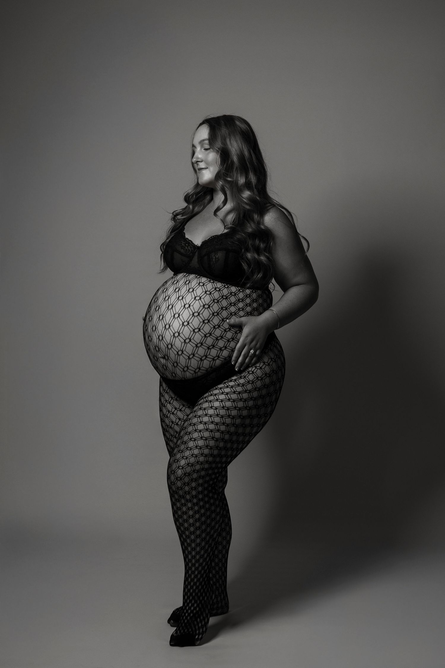 Black and white maternity photo of a smiling pregnant woman in lace bodysuit and patterned tights, posing in studio.