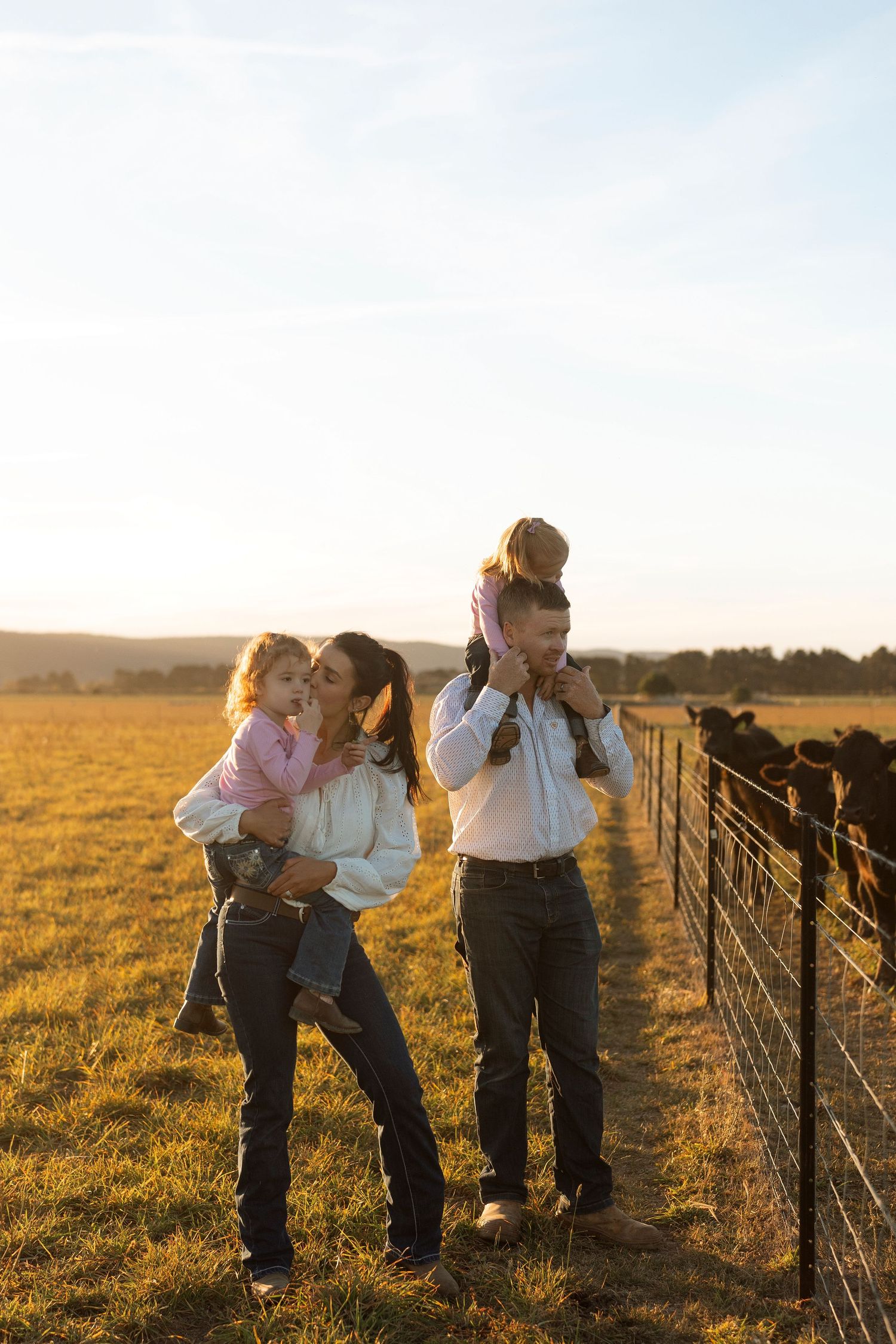 Family of four enjoying golden hour sunset on a farm, parents carrying young children near a cattle fence in a green field.