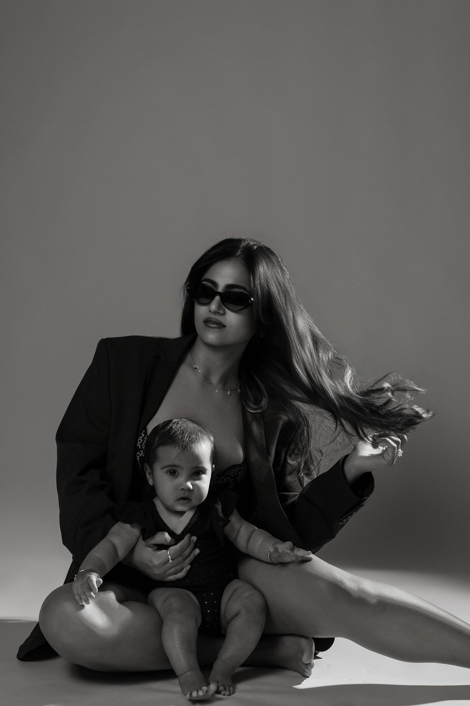 Black and white photo of a stylish woman in sunglasses and blazer sitting with a baby in a studio setting.