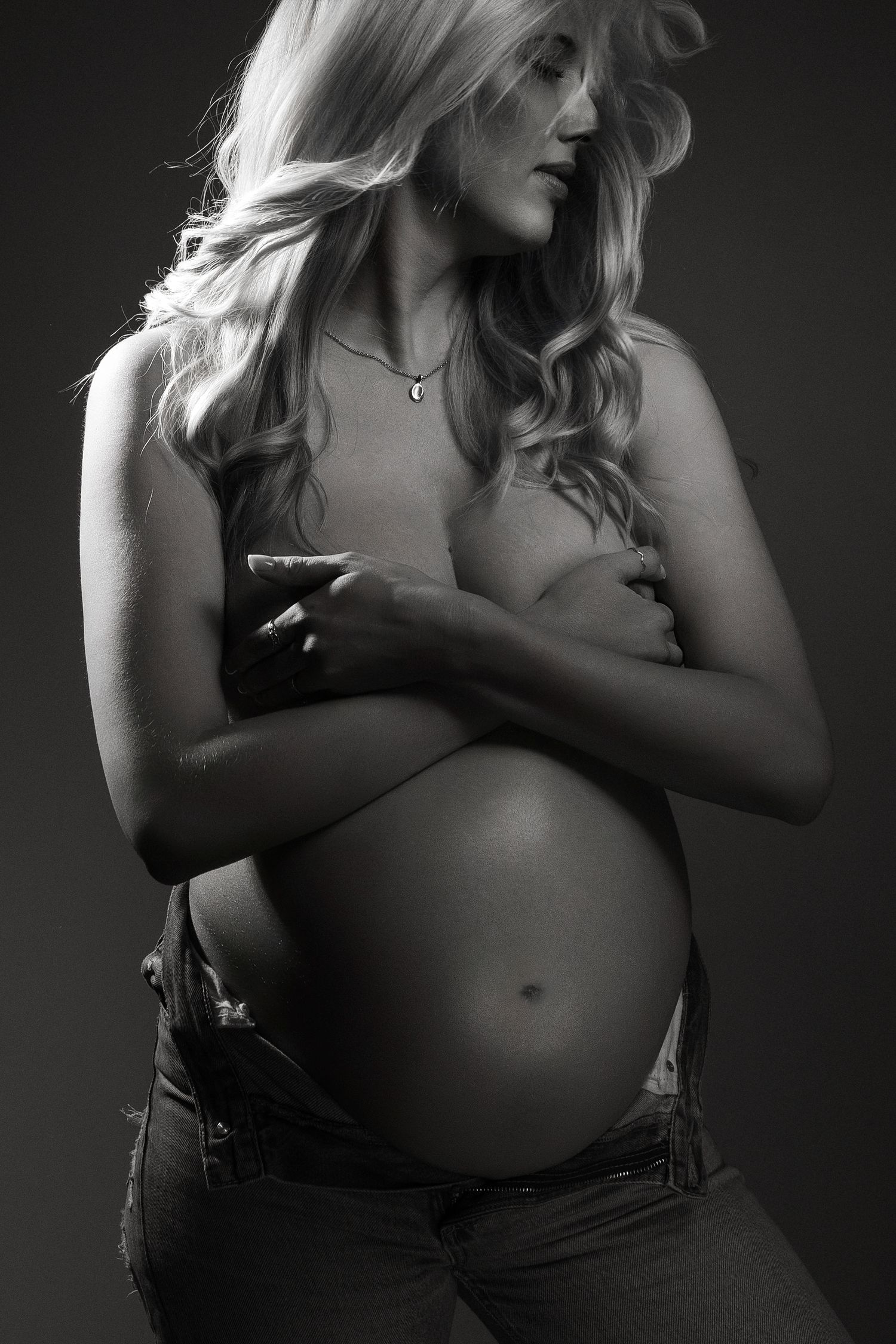 Black and white portrait of a pregnant woman with long blonde hair cradling her bare baby bump in studio lighting.