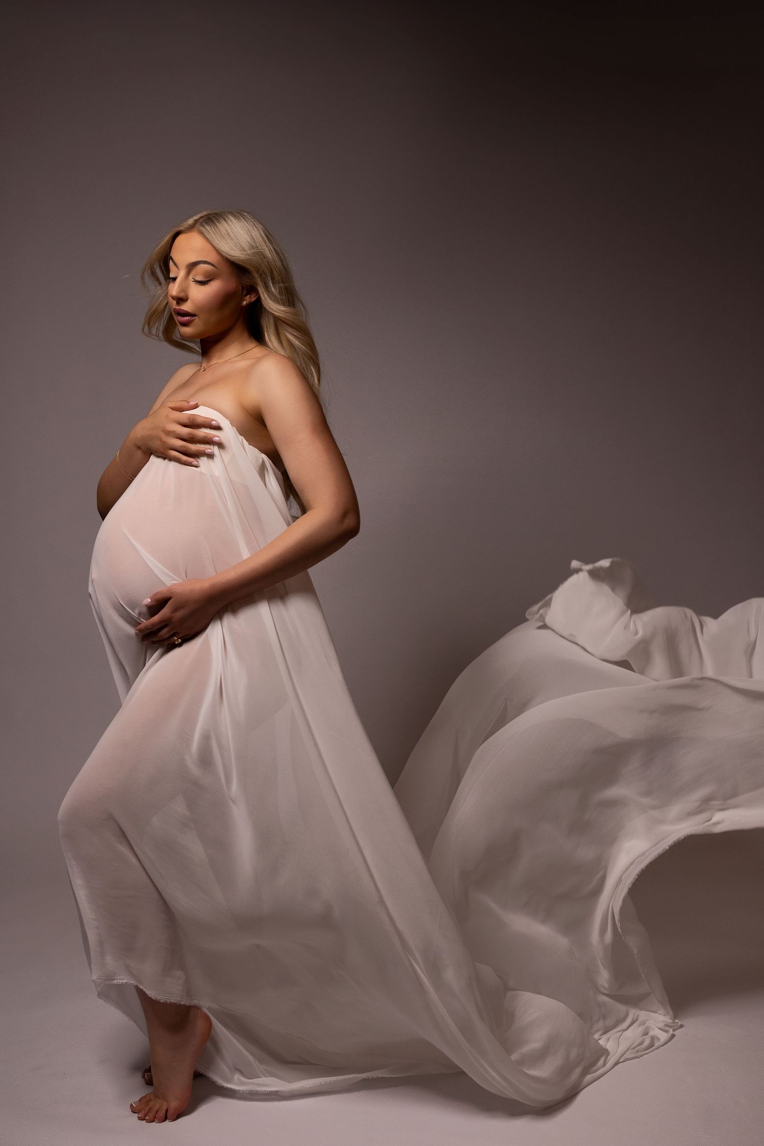 Pregnant woman draped in flowing white fabric, cradling her baby bump in a serene studio maternity photo.