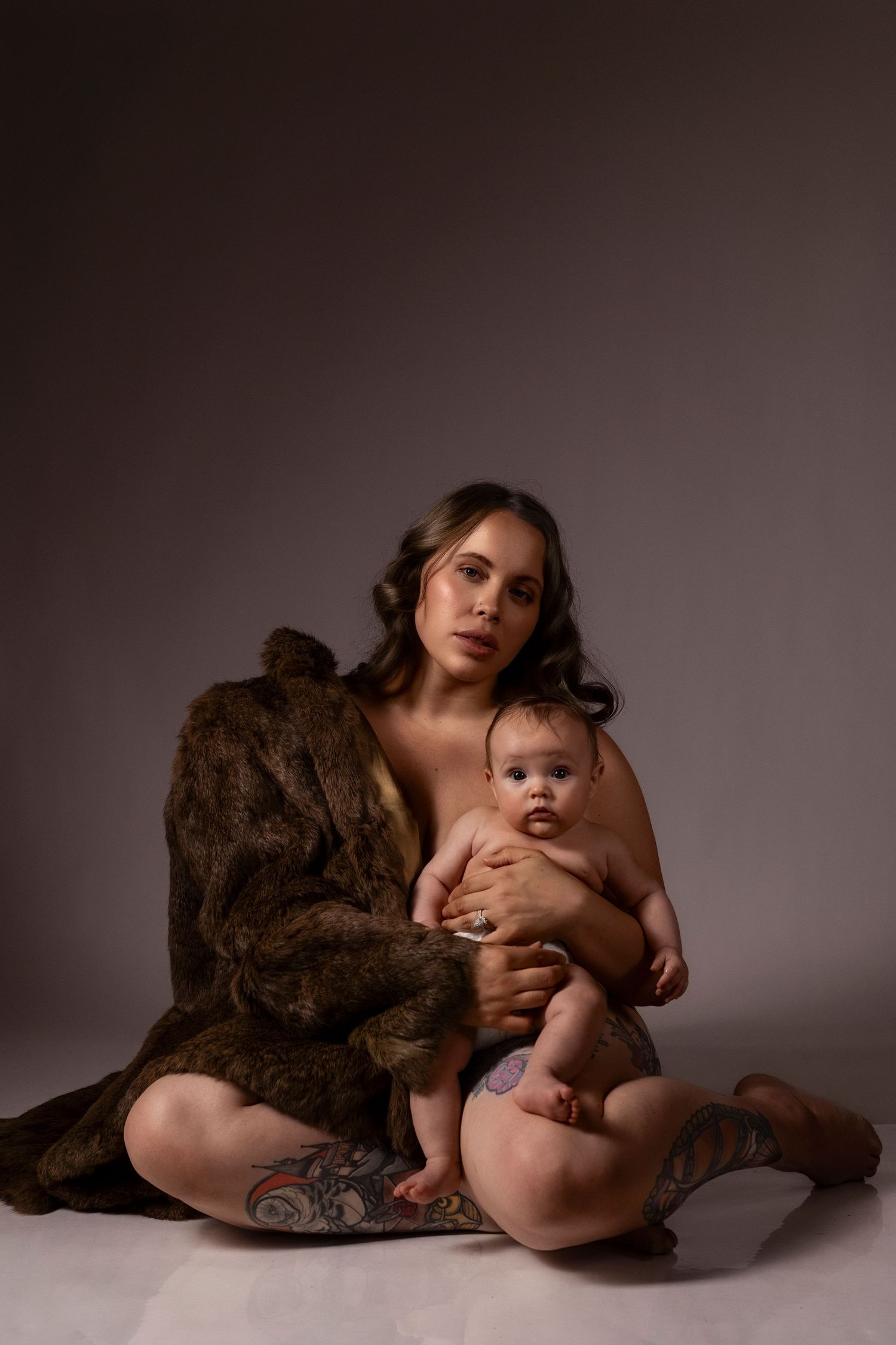 Mother and baby pose artistically on floor wearing brown fur coat in dramatic studio lighting.