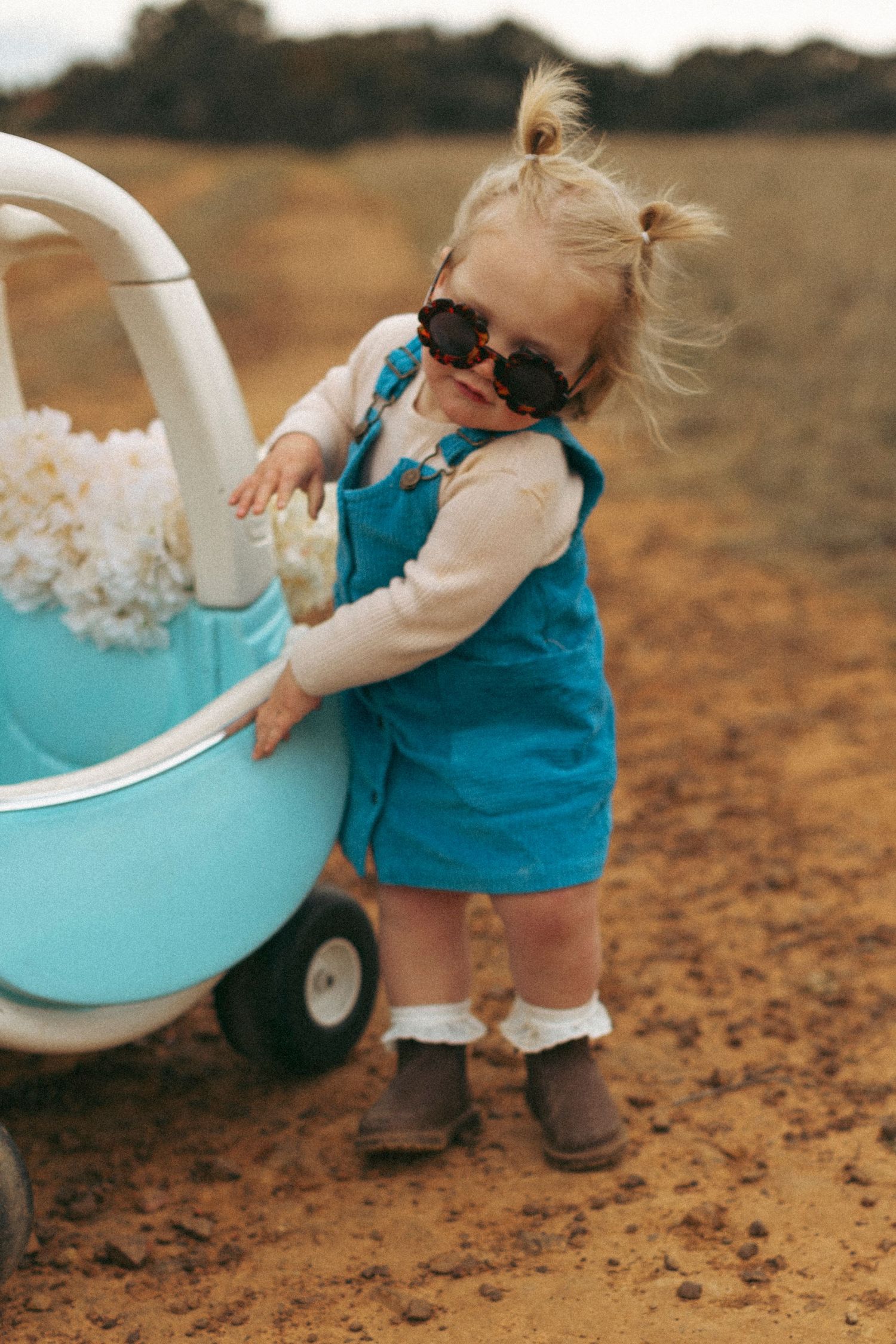 Small figure in blue dress playing with a turquoise toy wagon on a textured ground.