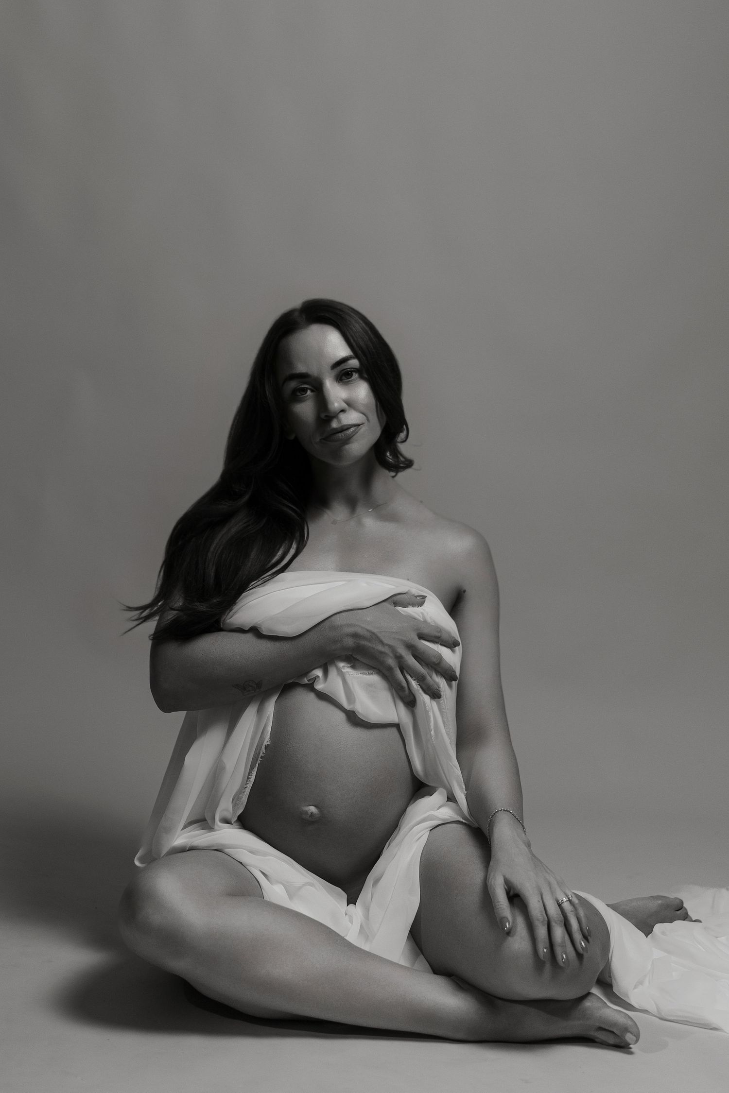 Black and white maternity photo of a woman sitting on the floor, draped in white fabric, cradling her pregnant belly.