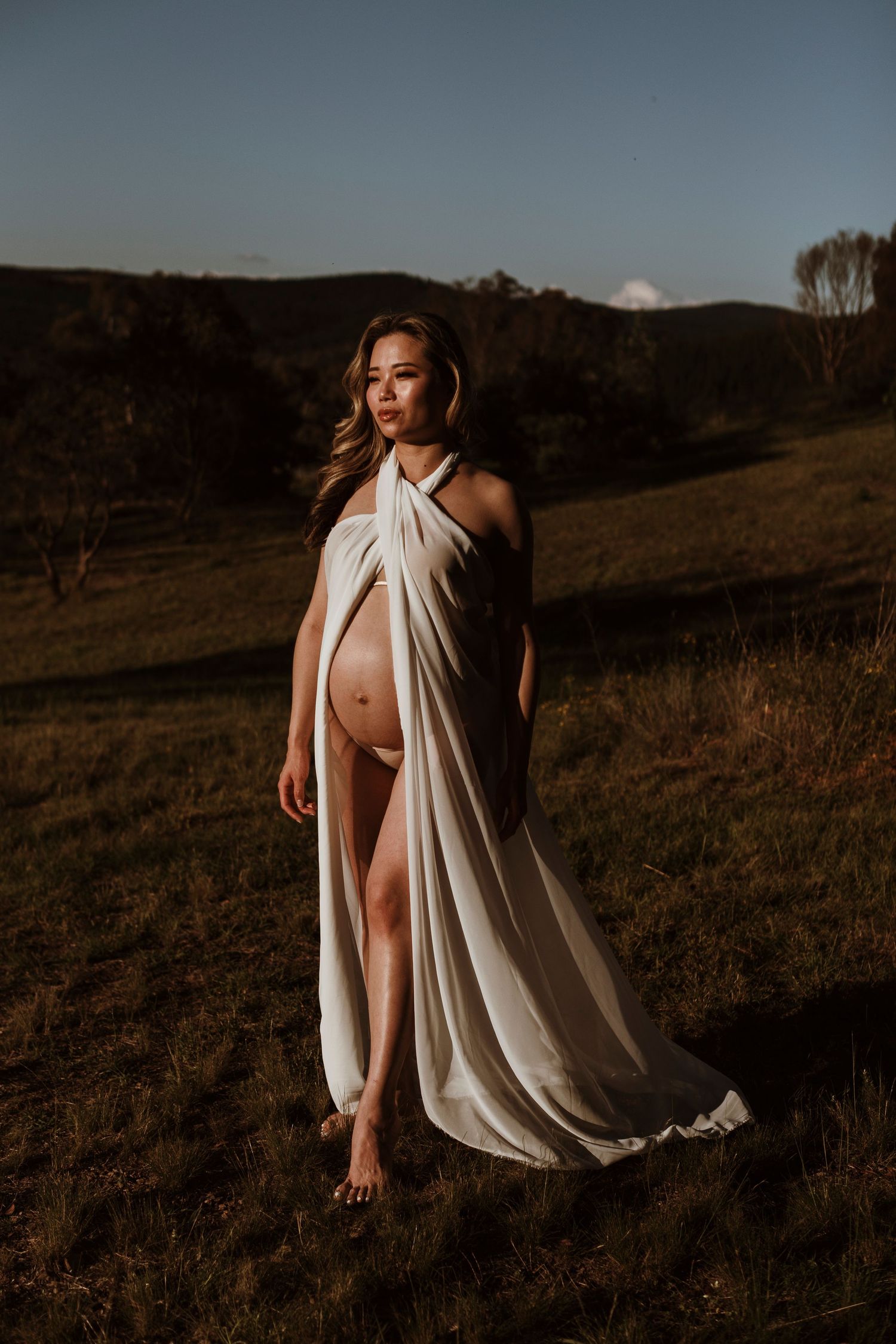 Artistic outdoor portrait with flowing white fabric against a mountainous landscape at sunset.