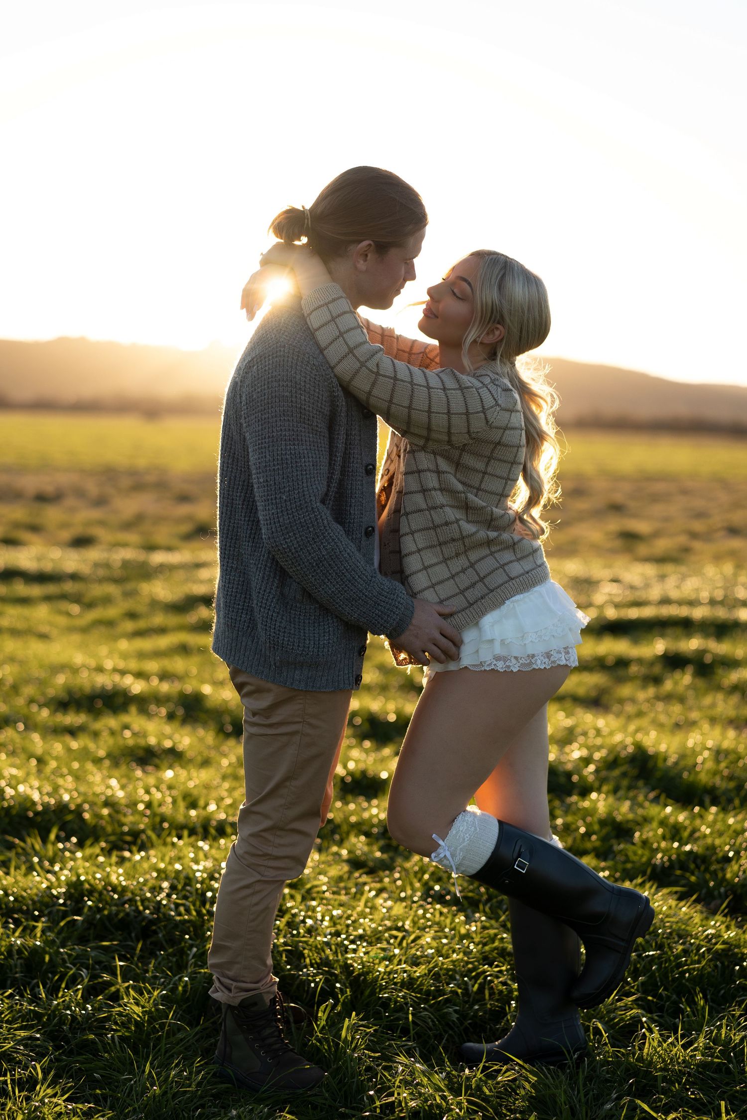 Couple embracing romantically in a sunlit green field at golden hour, woman wearing boots and white skirt.