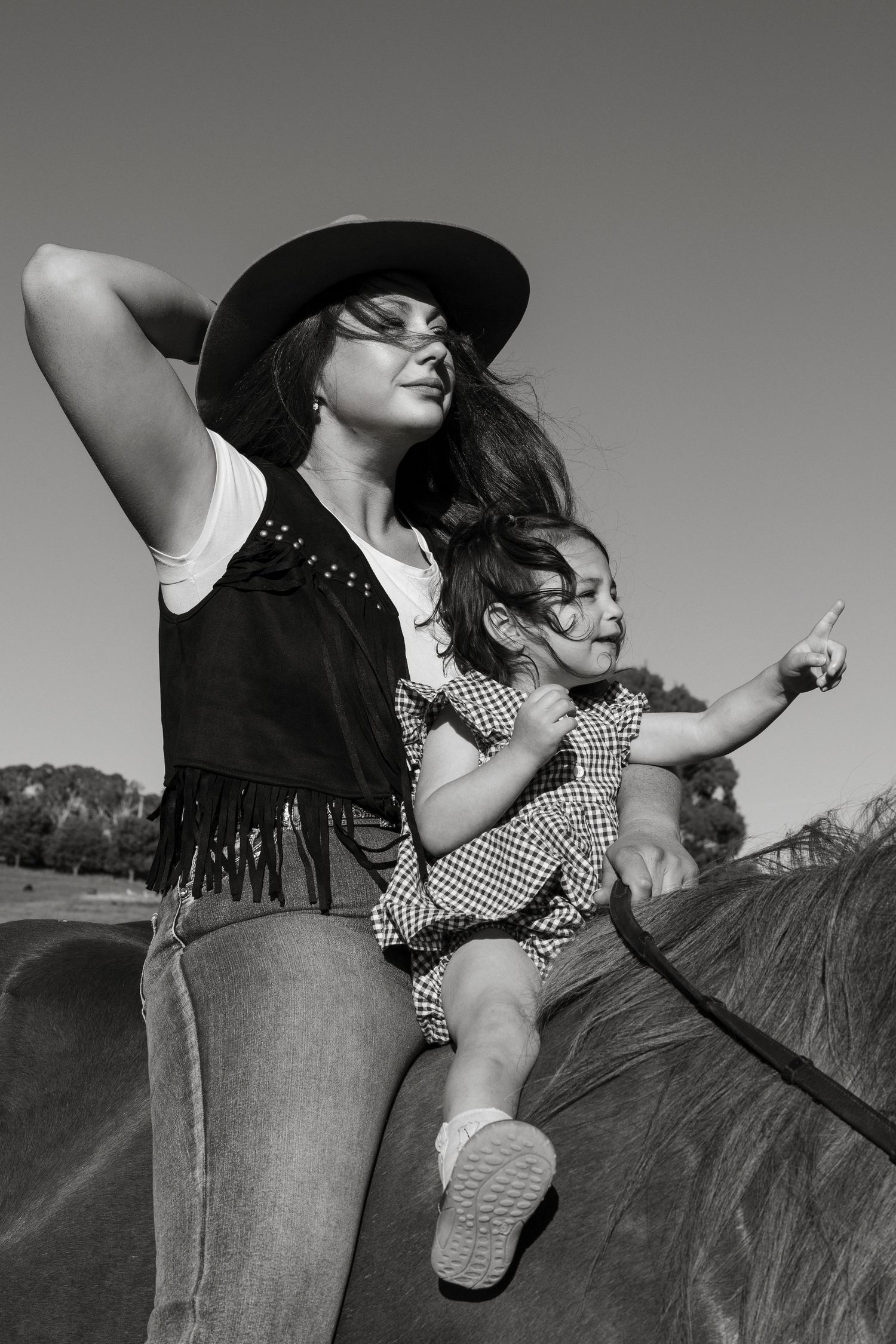 A black and white photo of two people riding horseback, with one wearing a hat and vest while the other wears polka dots.
