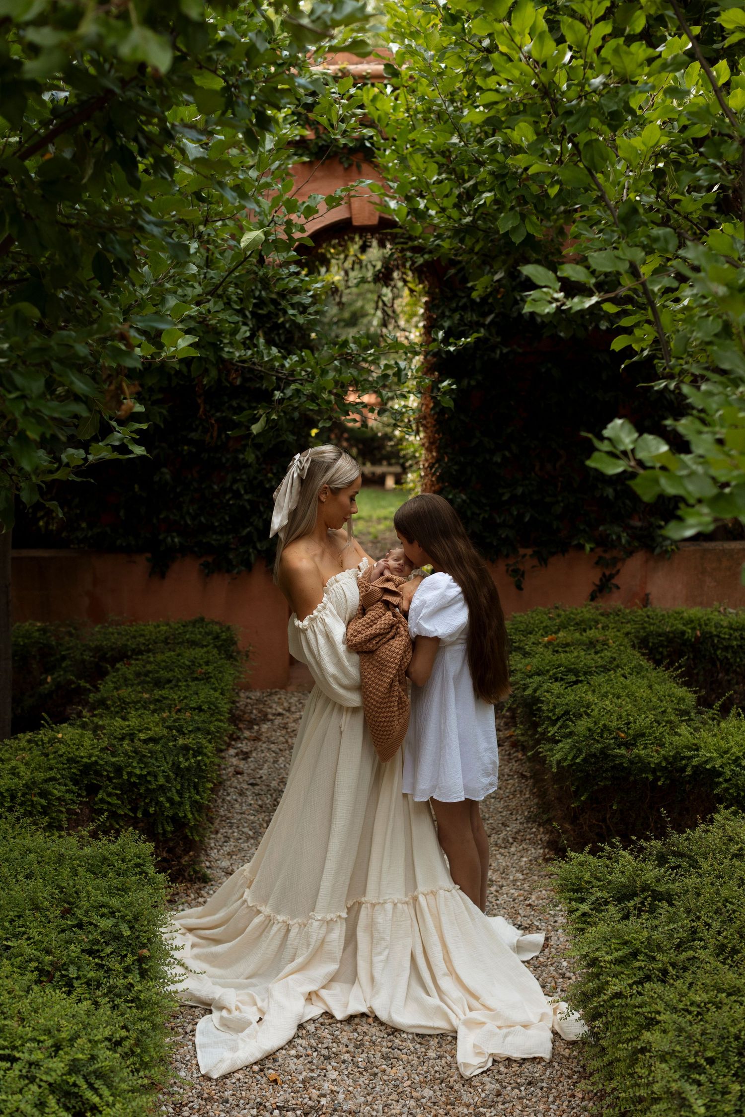 Mother and child in flowing white dresses embrace in lush garden setting.