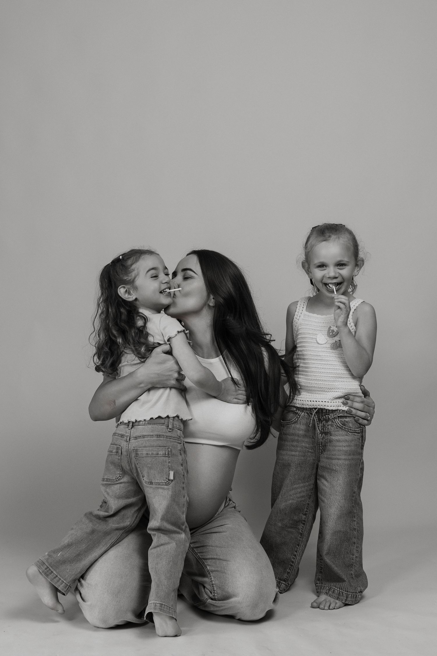 Black and white photo of a pregnant woman kneeling and kissing her daughter while another young girl smiles nearby.