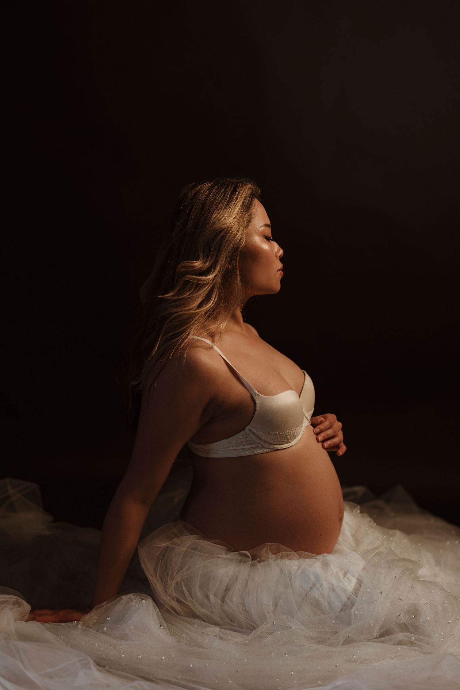 Maternity portrait with person in white lingerie sitting on tulle fabric.