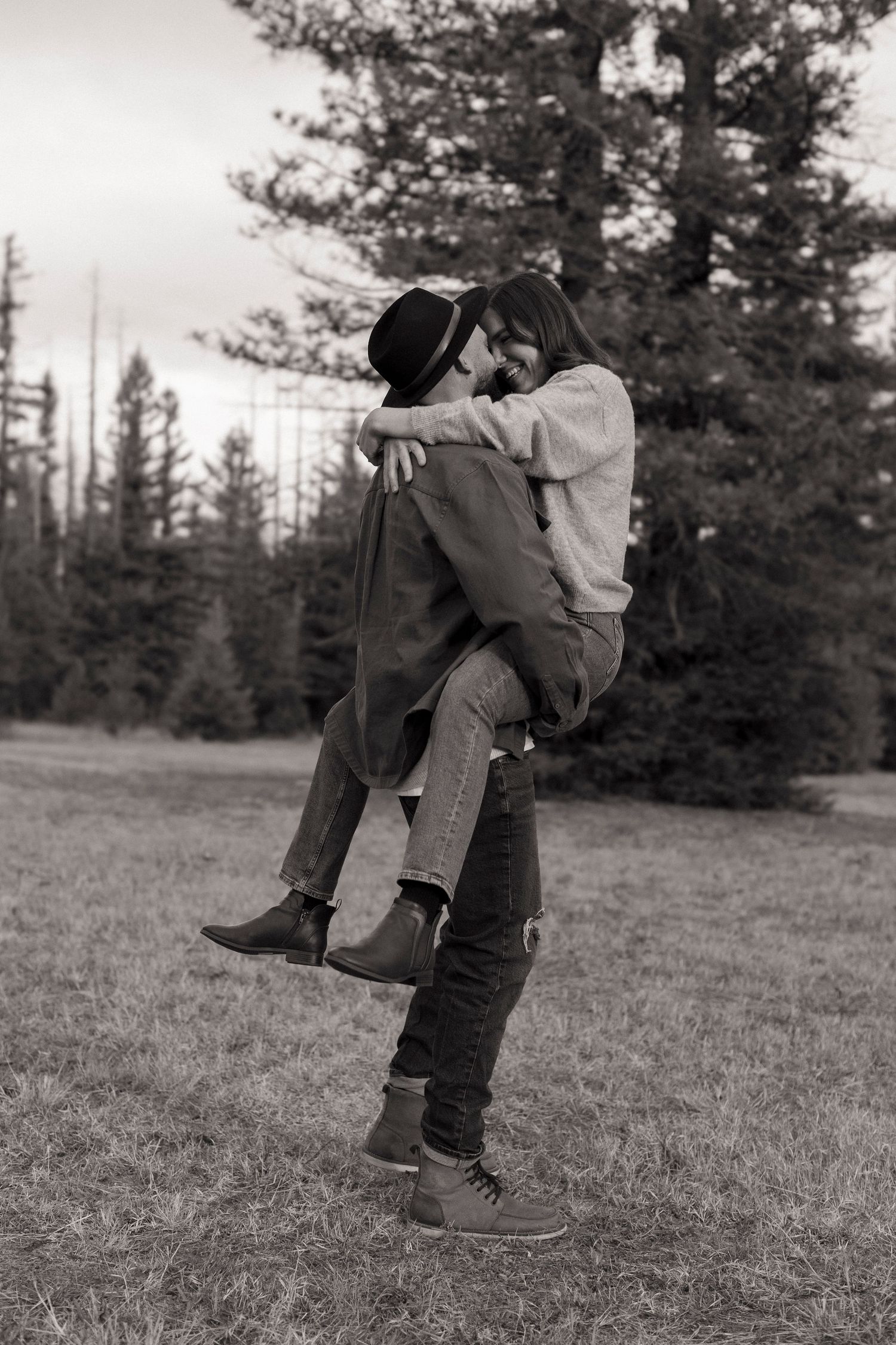Black and white outdoor photo of a romantic embrace between two people in a field with evergreen trees in background.