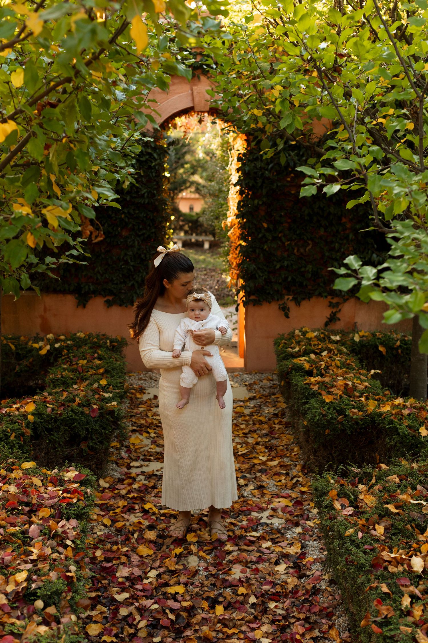 A tender moment captured in autumn sunlight as fallen leaves create a golden carpet beneath trees in a garden setting.