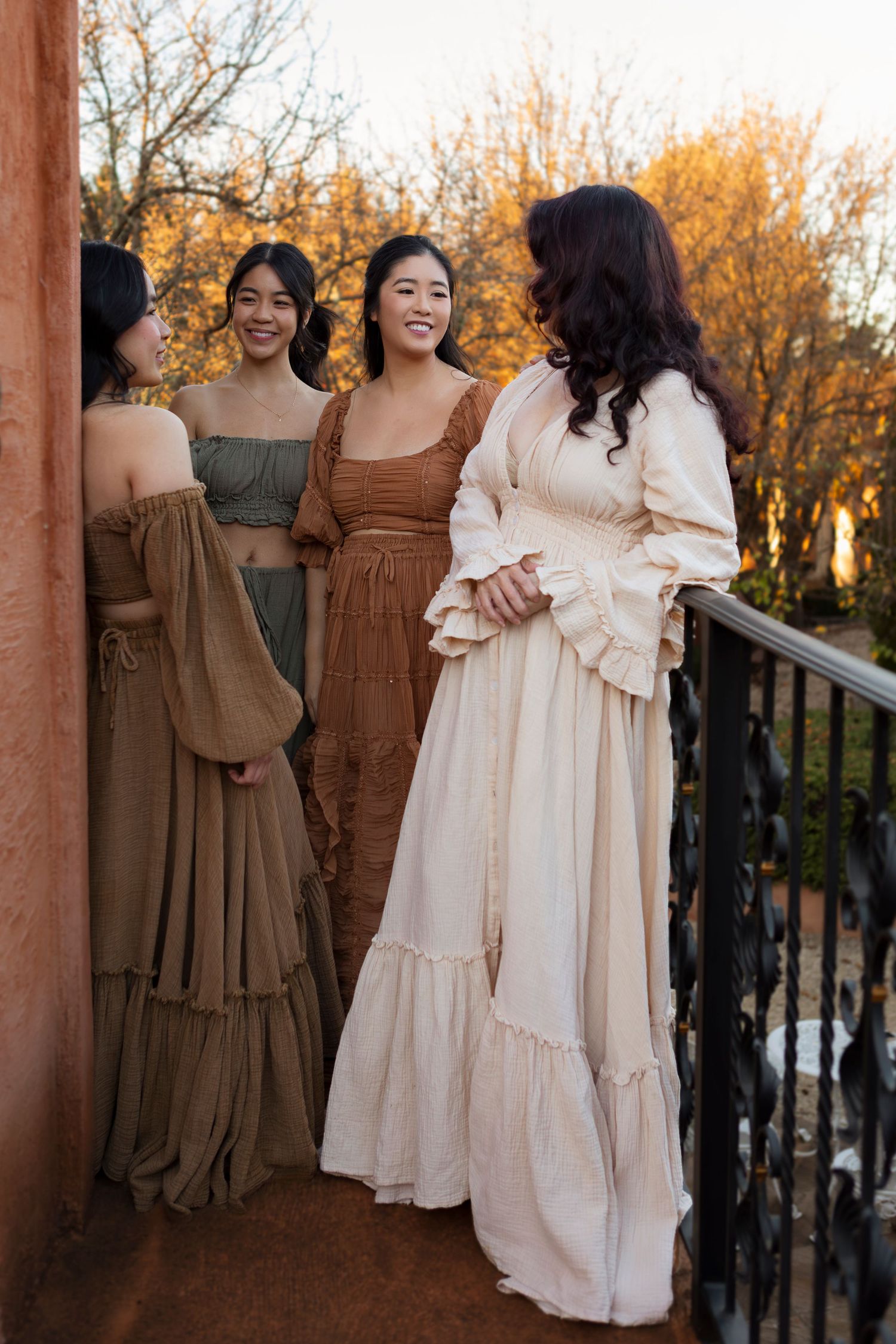 A group stands together wearing coordinated brown and cream colored dresses in an outdoor autumn setting at sunset.