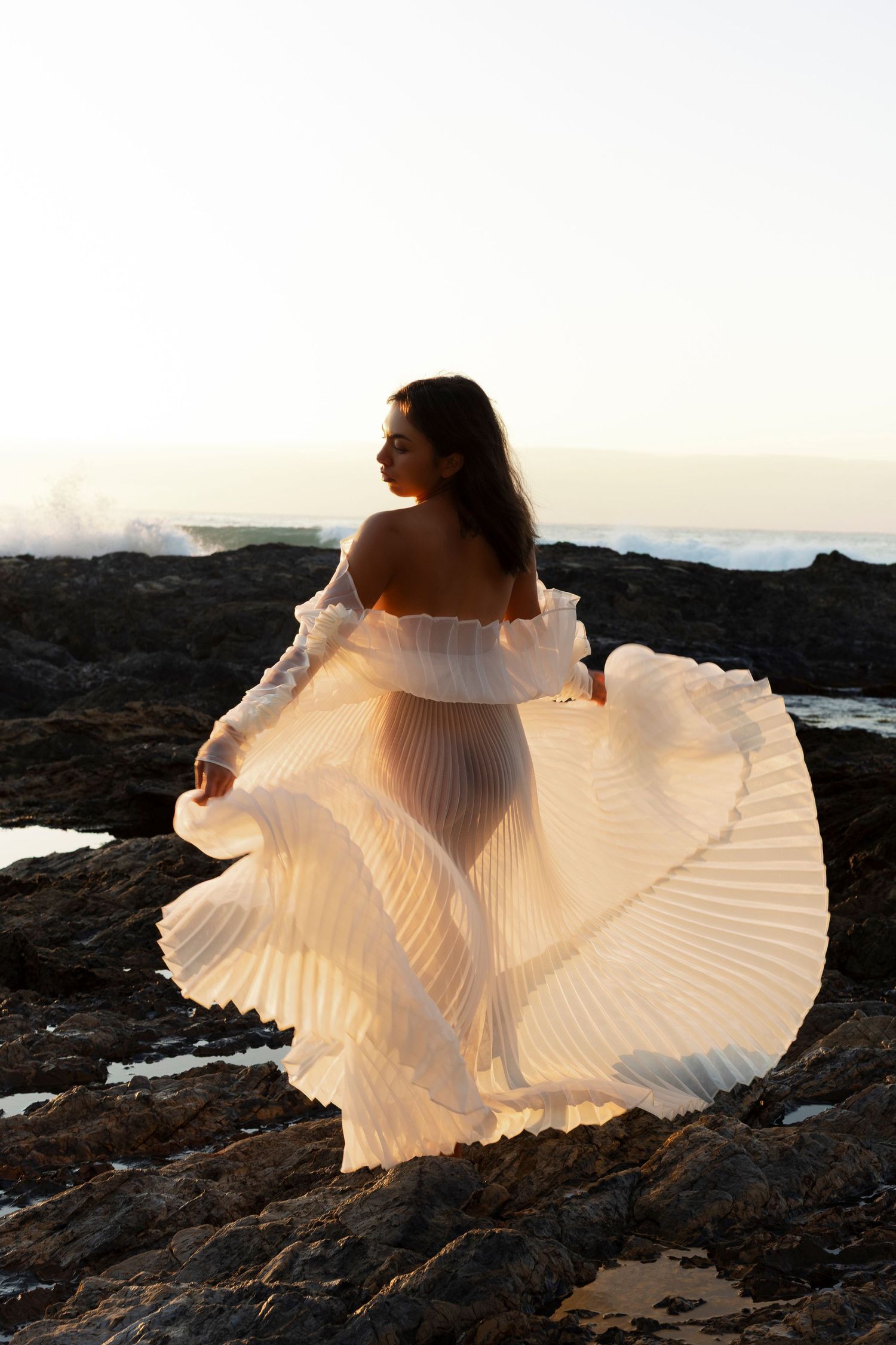 Figure in flowing white pleated dress posing gracefully on rocky coastal landscape at sunset.